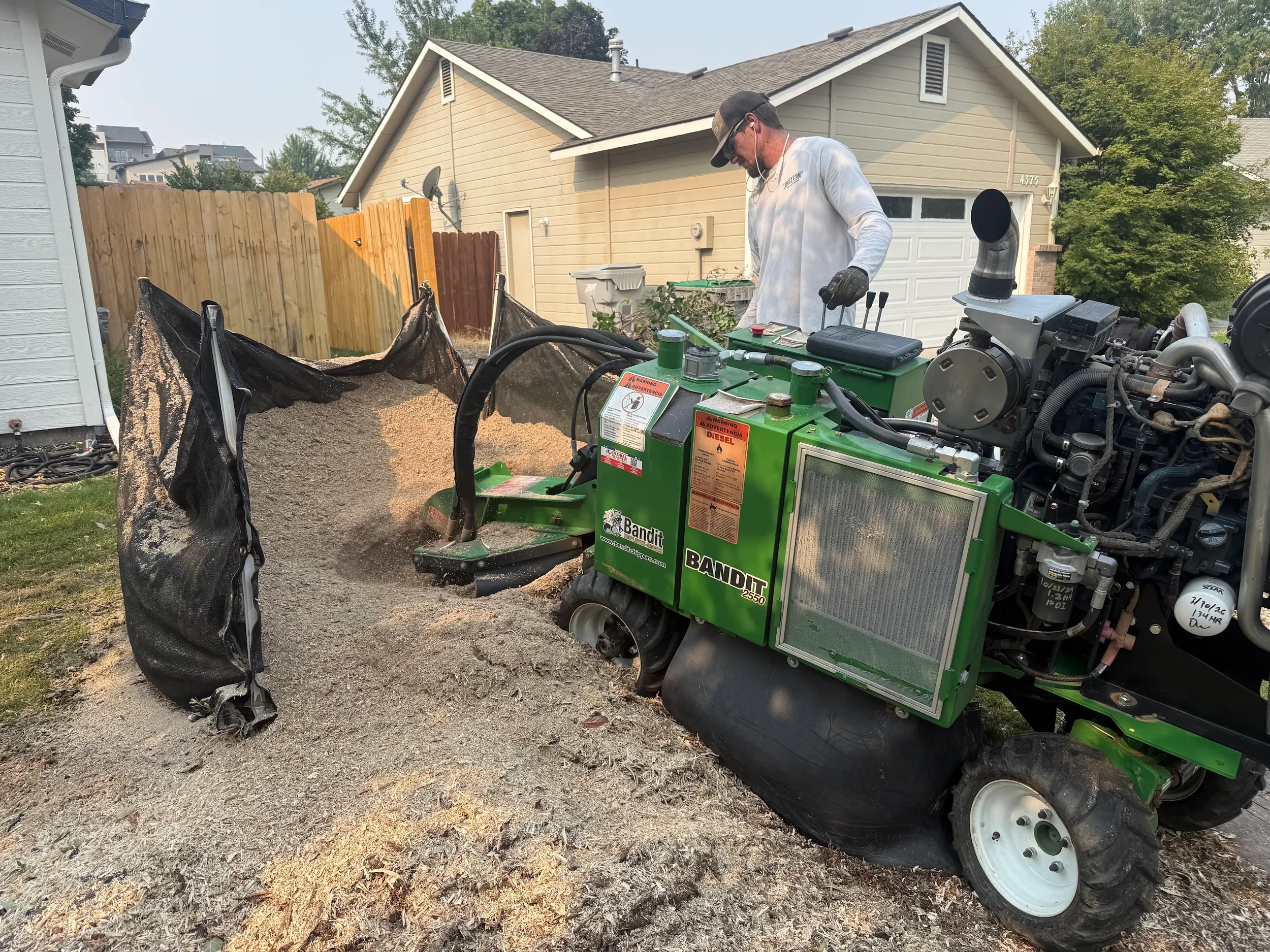 Man operating a green wood chipper in a backyard, feeding branches into it with a pile of wood chips on the ground, houses, and trees in the background.
