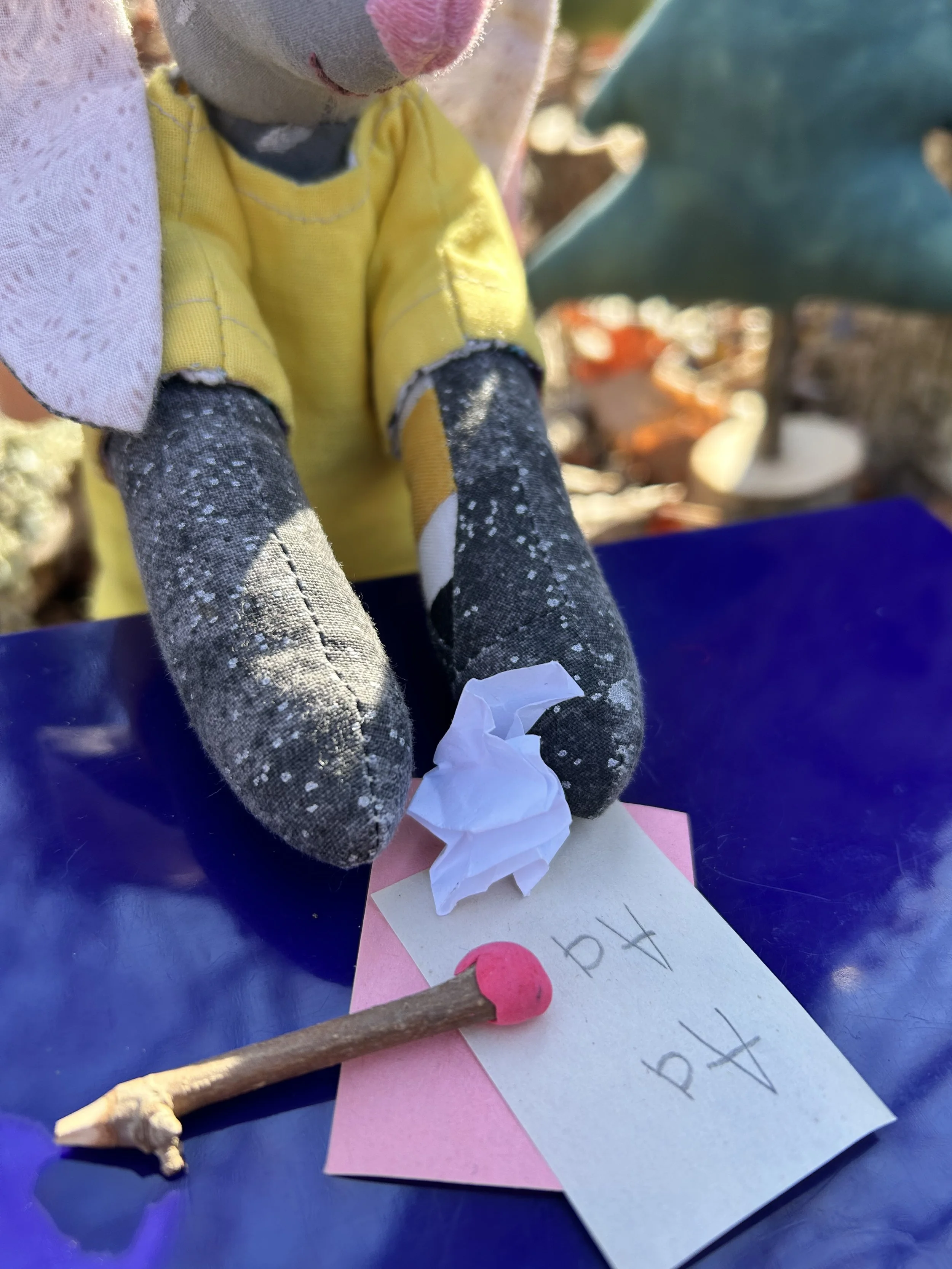 A close-up of a handmade doll with a gray face, pink nose, and black hands, wearing a yellow shirt. The doll is holding a white paper with handwritten notes and a pink star-shaped paper underneath. There is a wooden stick with a pink tip and a truncated piece of white clay or chalk on the blue surface.