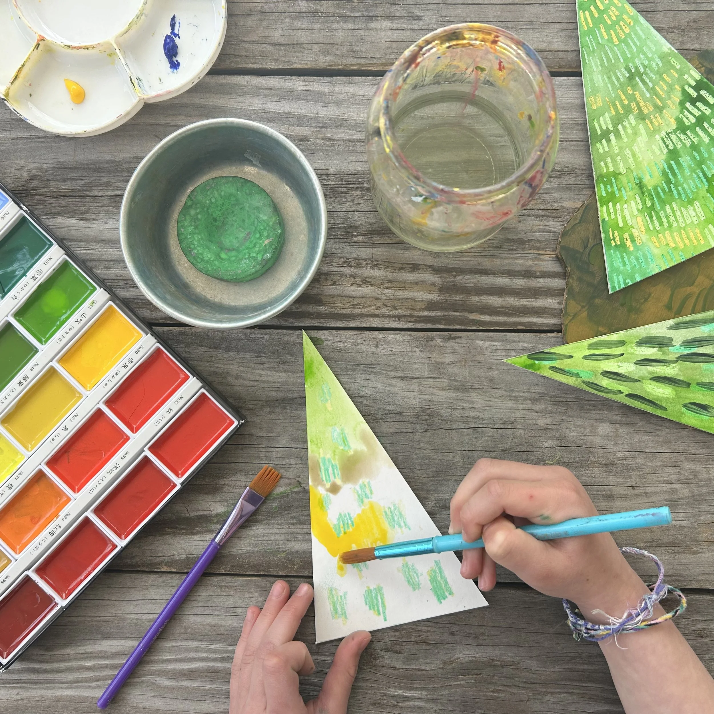 Person's hands painting with markers on a piece of paper, surrounded by watercolor paints, a glass jar, and a plastic palette on a wooden surface.