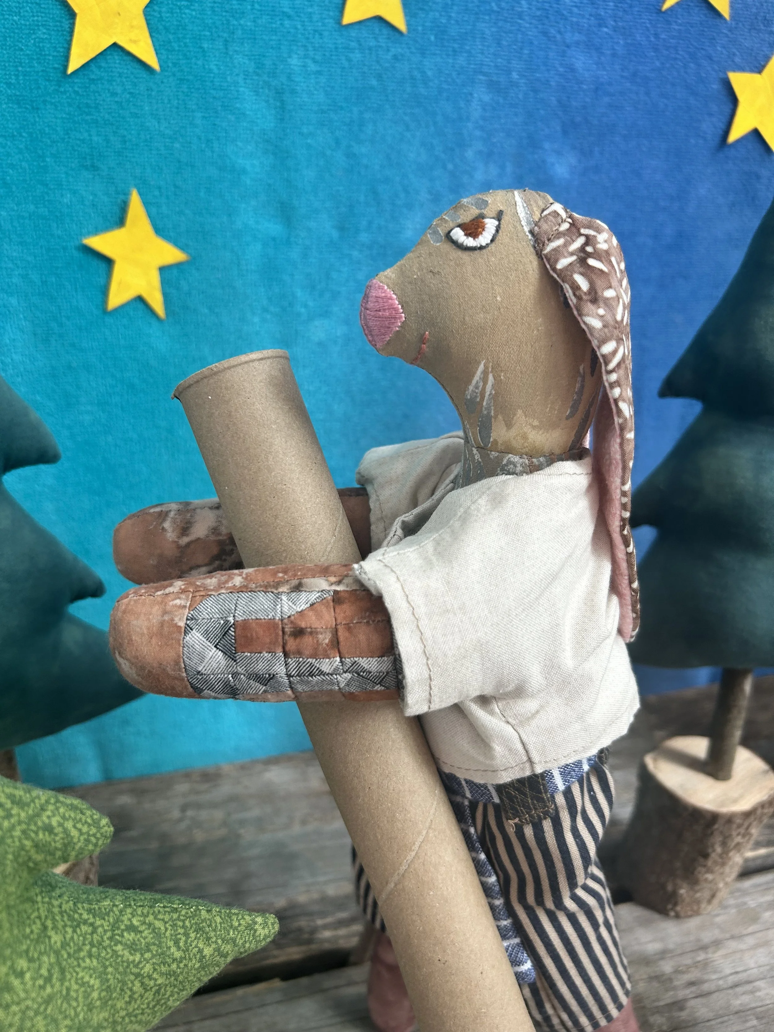 A handmade fabric puppet with a beige face, brown eyes, pink lips, and brown ears with white spots. The puppet wears a white shirt and striped brown and white pants, holding a cardboard tube, in front of a blue backdrop decorated with yellow stars.
