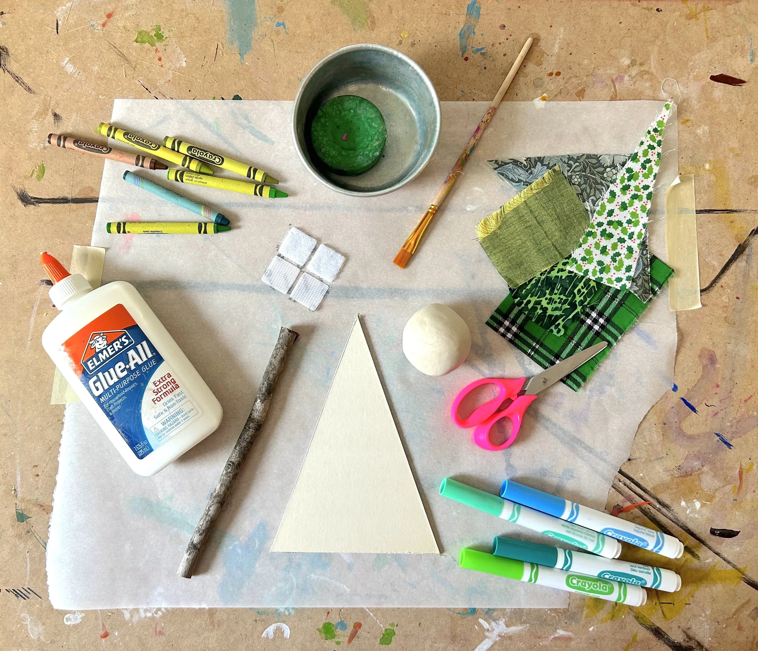 A tabletop with art supplies, including markers, glue, paper, scissors, a small cup with green paint or glue, fabric pieces, small white foam squares, and a glue stick. The surface is covered with paint splatters and marks.