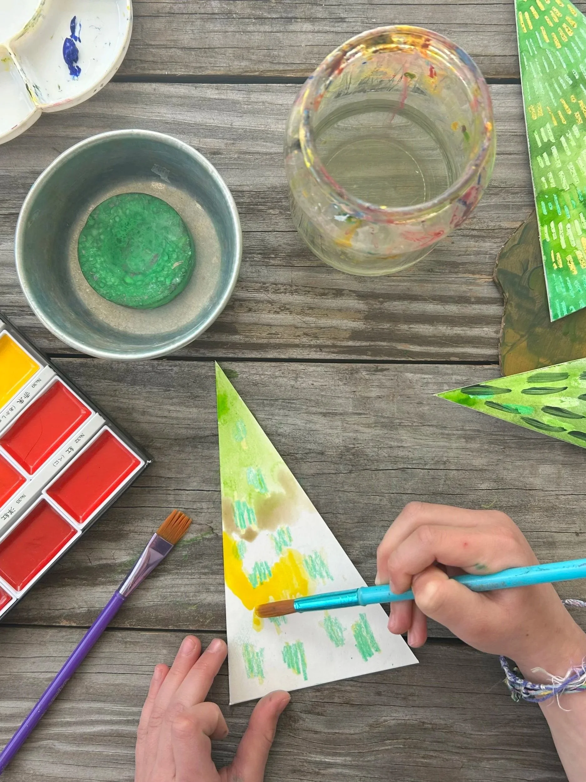 People painting a tree shape using watercolors and brushes. The scene includes green and yellow colors, with some blue strokes. Art supplies such as watercolor palette, brushes, and a water jar are visible on a wooden surface.