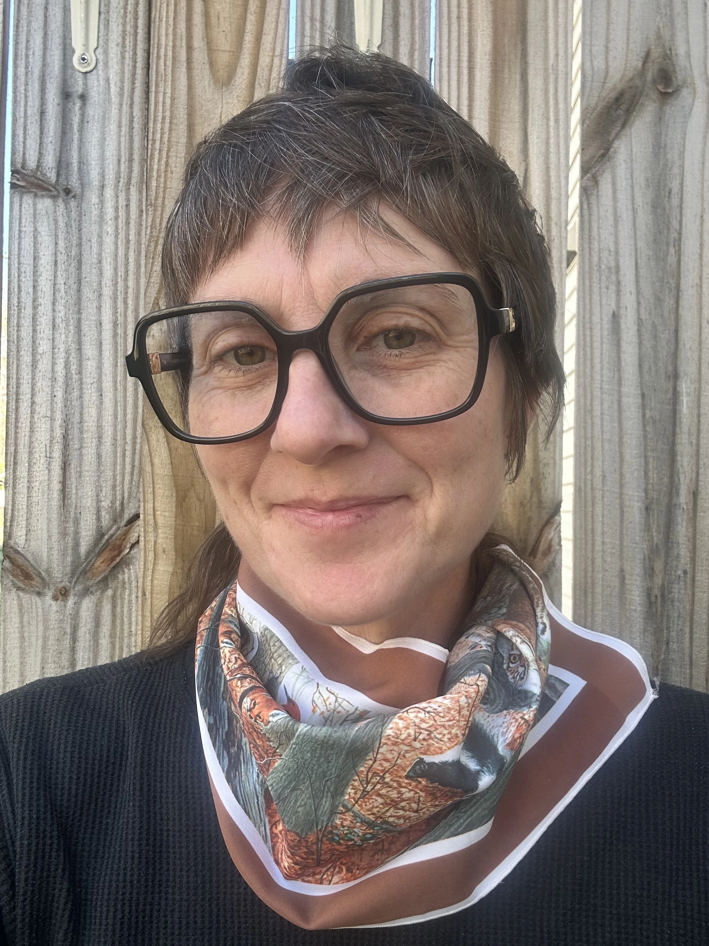Close-up of a woman with short, dark hair and glasses, wearing a scarf with a nature scene, standing in front of a wooden fence.