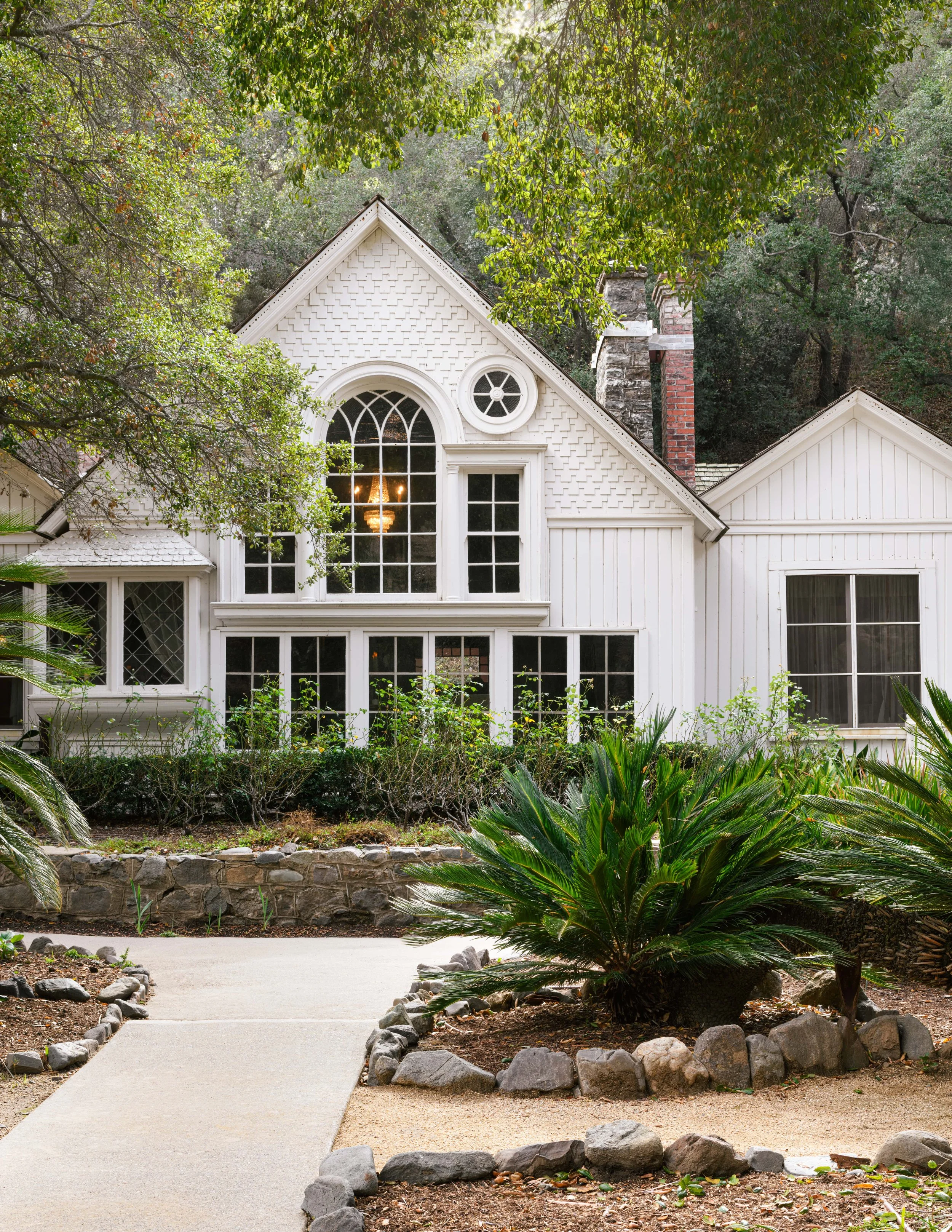 White house with large arched window and smaller round window, surrounded by greenery, with a garden and stone border in the foreground.