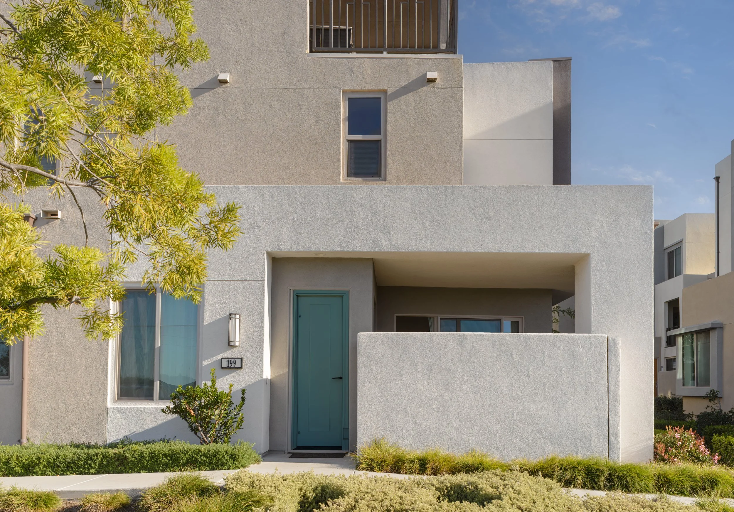Modern two-story house with a teal front door, white stucco walls, and surrounding green shrubbery and trees under a blue sky.