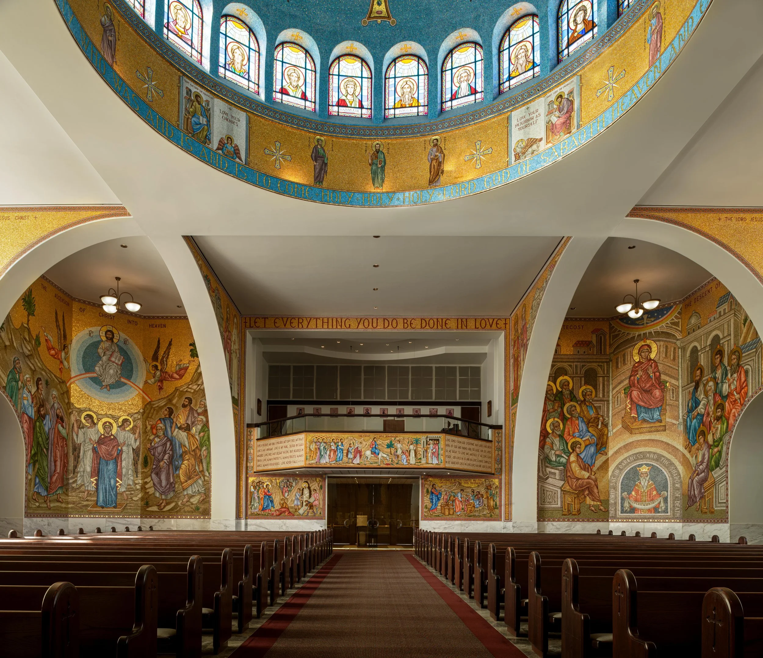 Interior of a church with stained glass windows on a dome ceiling and religious icons, murals, and gold accents on the walls.