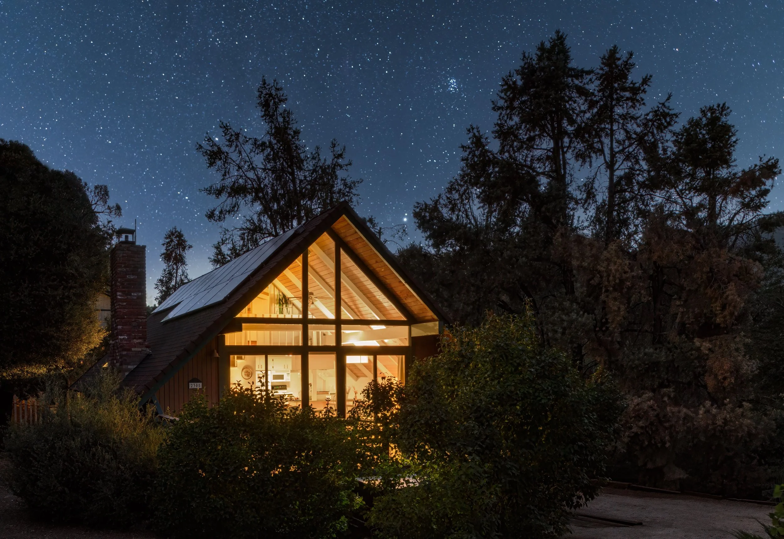 A house with large glass windows illuminated from the inside at night, surrounded by trees, under a starry sky.