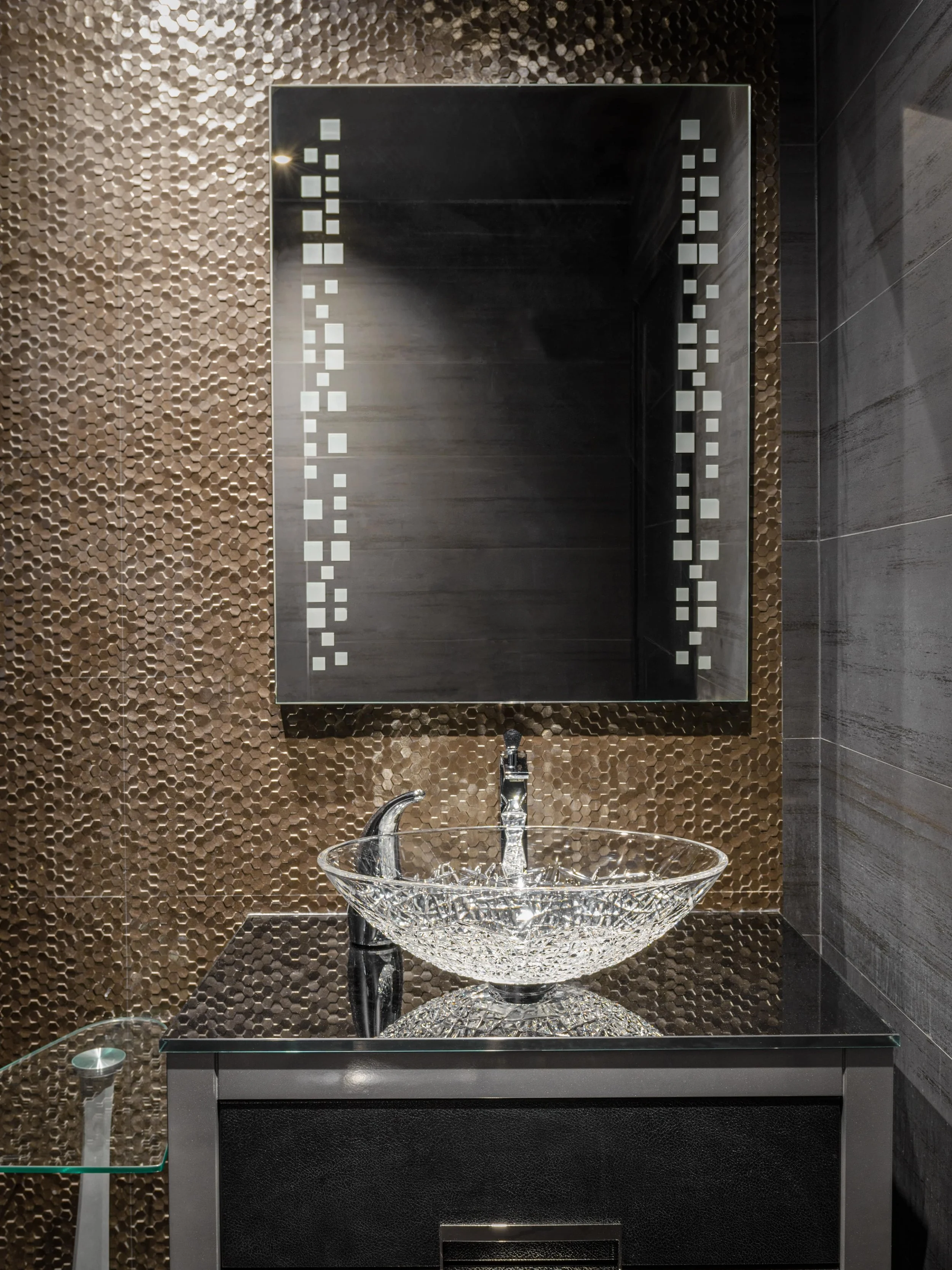 Bathroom vanity with a glass bowl sink, a large mirror with decorative border, and textured metallic brown and dark gray tiled walls.