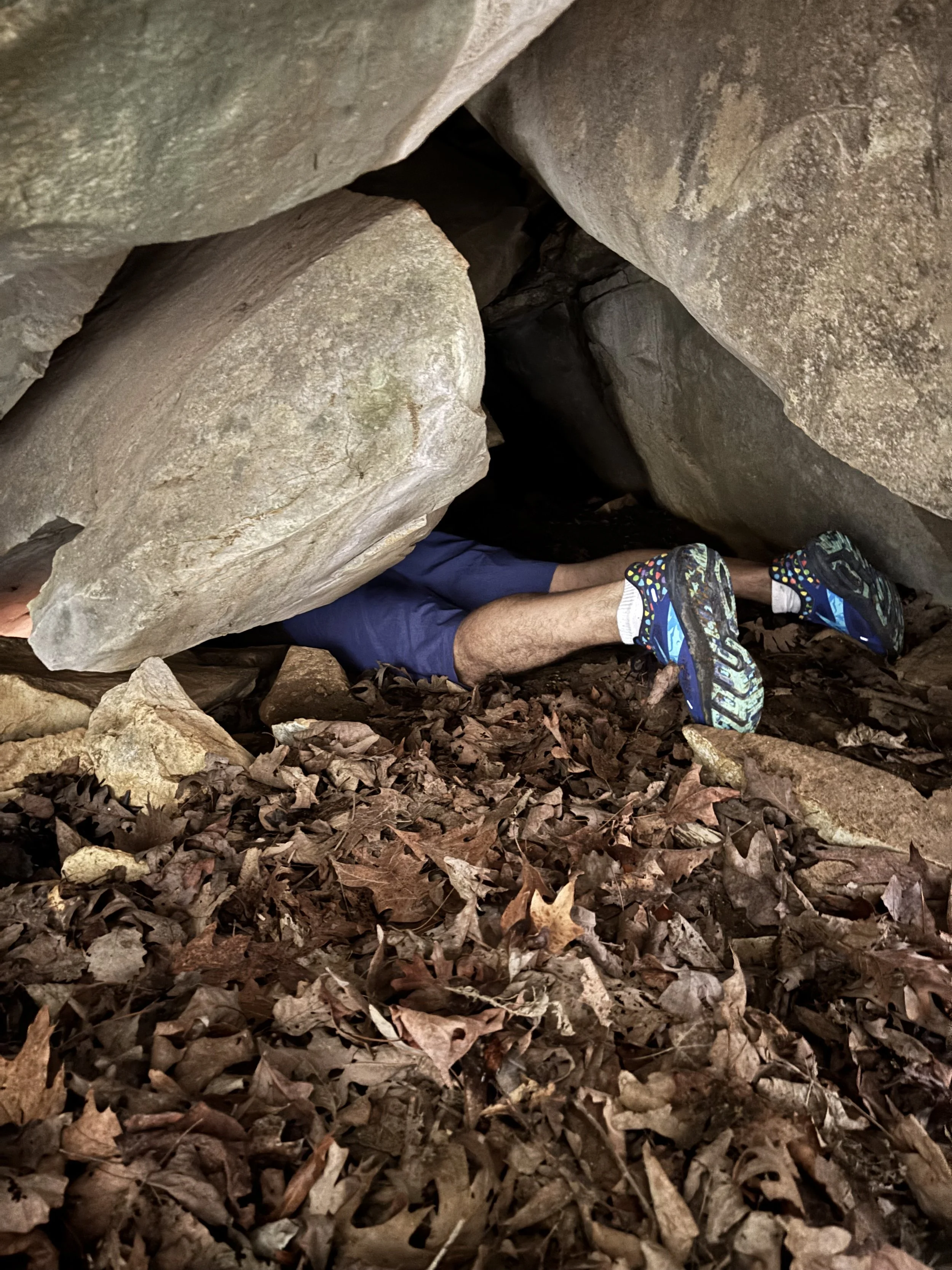 Person crawling through a small cave or crevice with large rocks above and dried leaves on the ground.