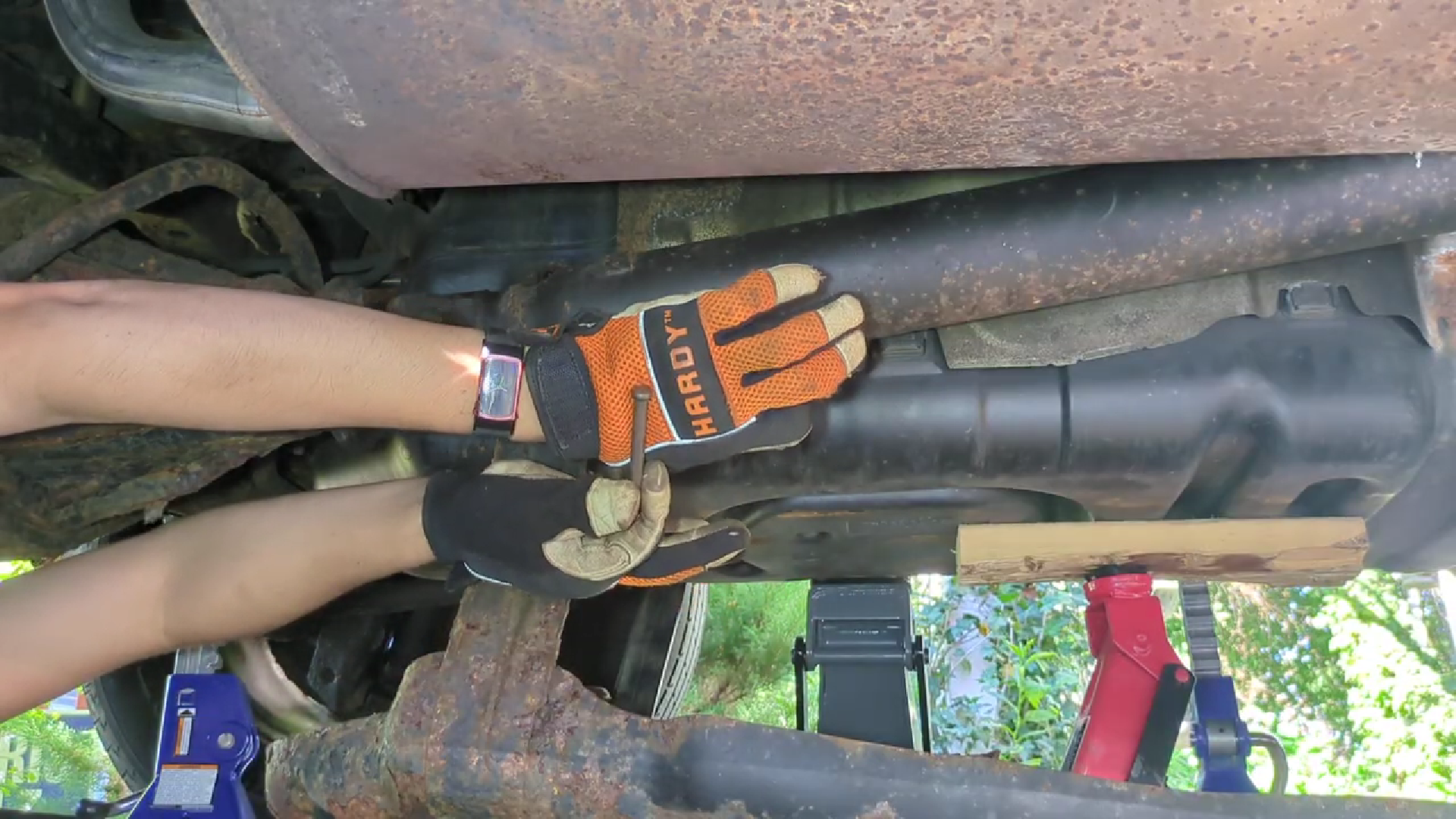 Under a car, that is supported by jackstands. Two jacks are visible, one is holding the gas tank up against the frame. Arms come from the left, with gloved hands holding a pin.