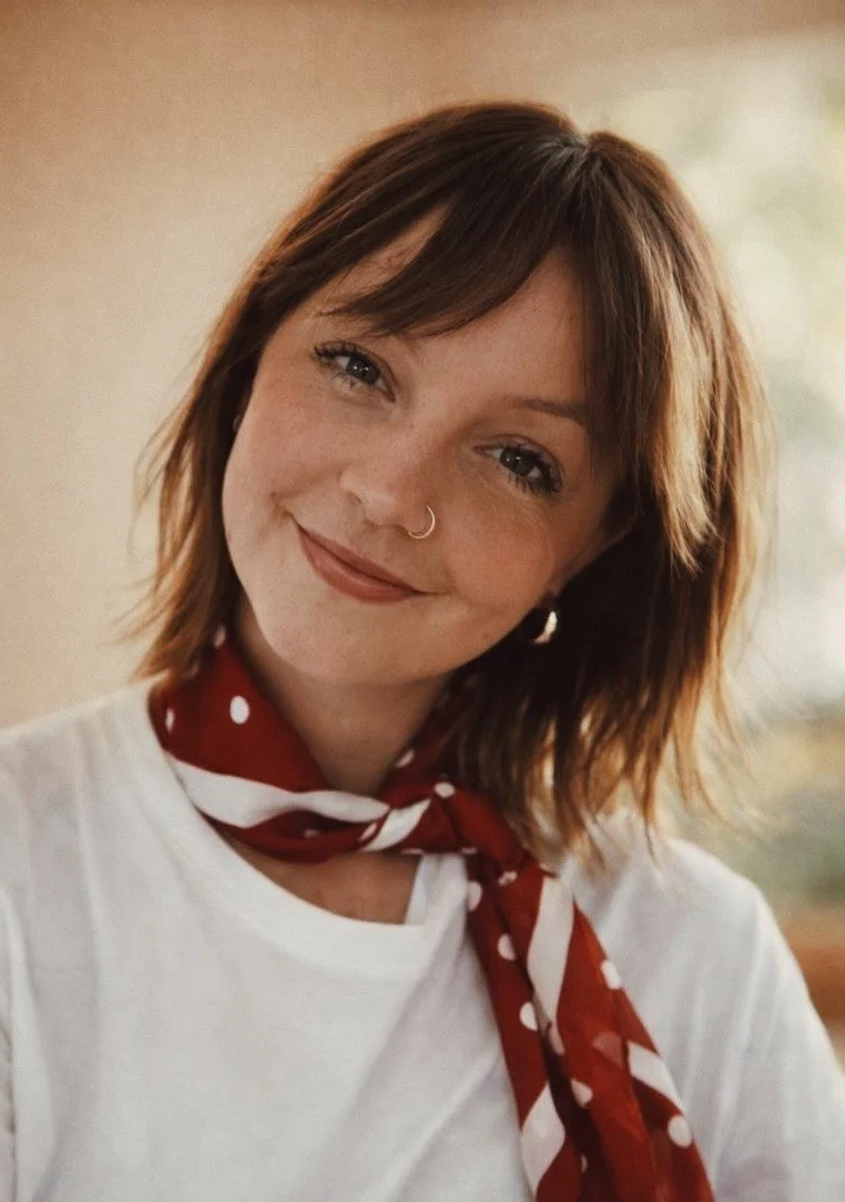 A smiling woman with shoulder-length brown hair, wearing a white shirt, a red polka dot scarf, and a nose ring.