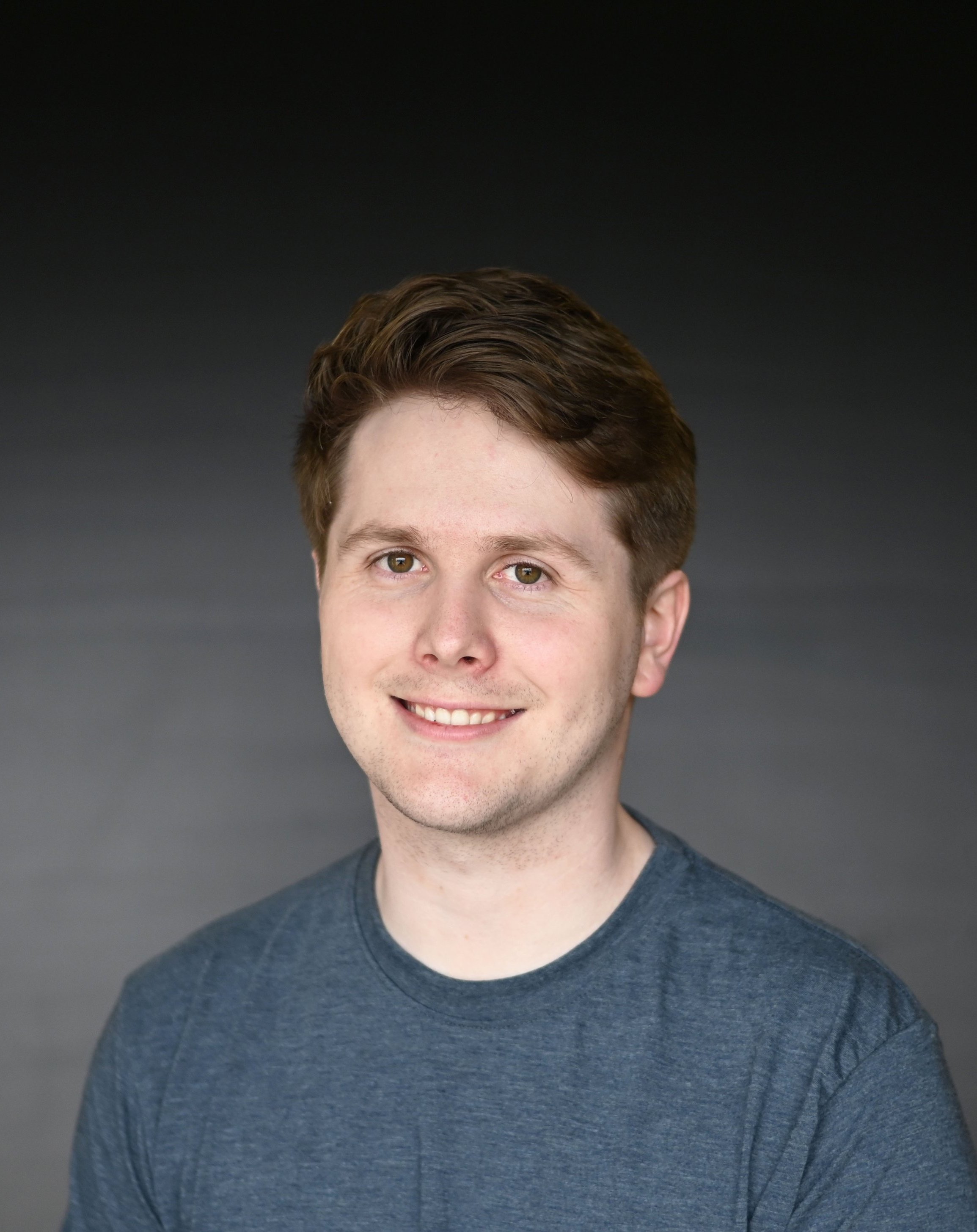Close-up of a young man with brown hair, smiling, wearing a dark gray T-shirt, against a dark gray background.