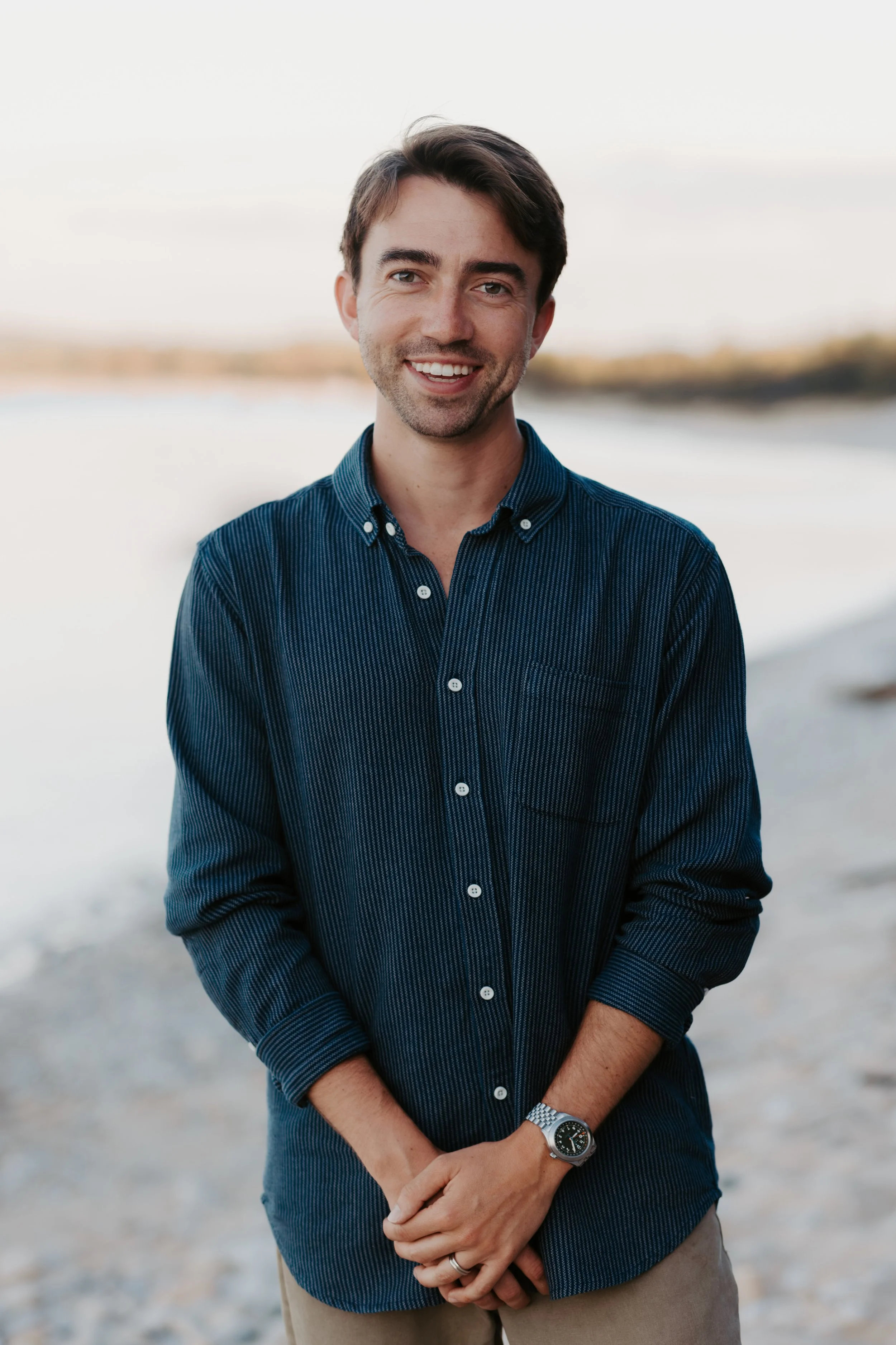 A young man with dark hair and a beard, wearing a dark blue button-up shirt, standing outdoors near a body of water, smiling at the camera.