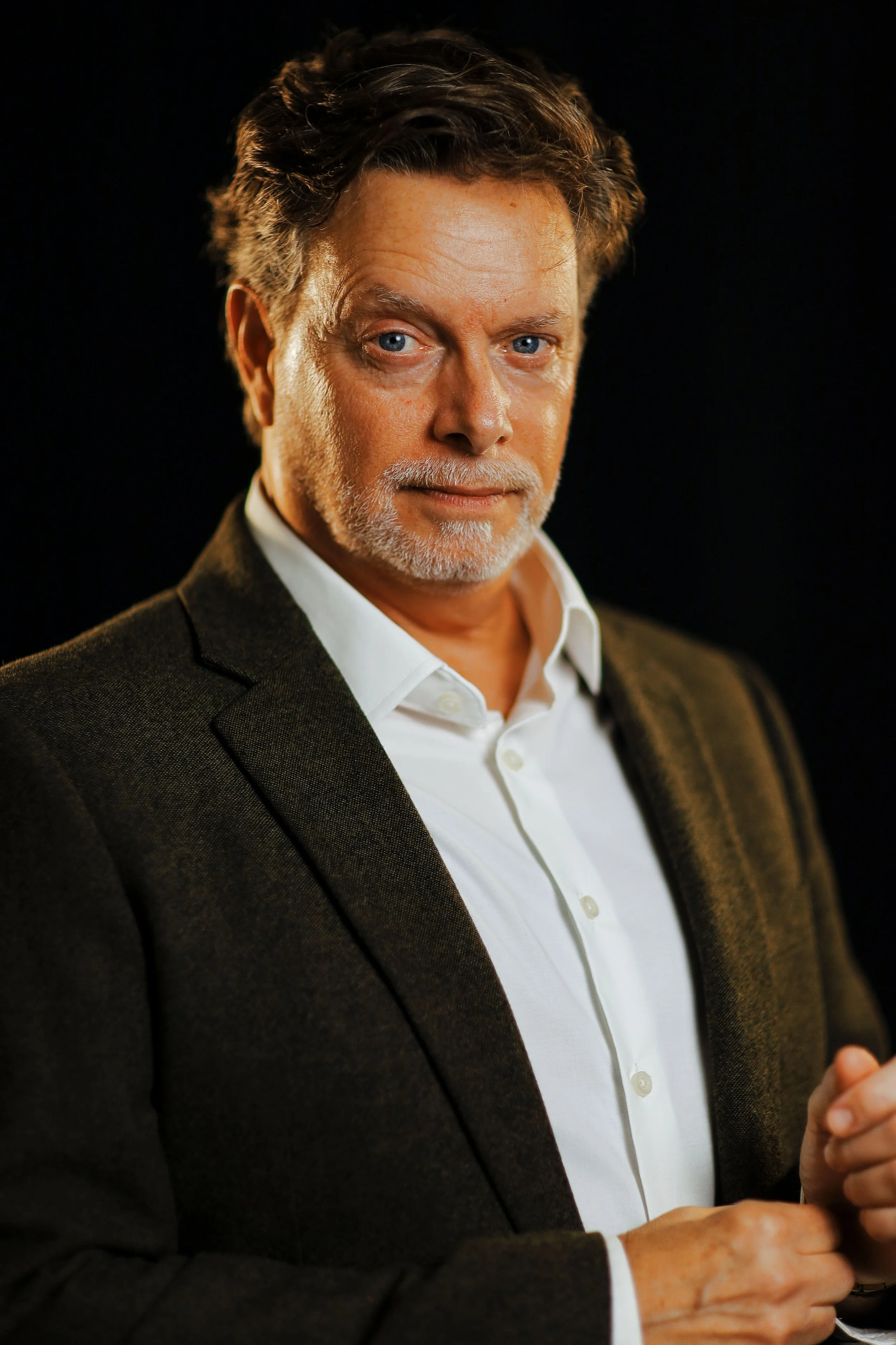 Close-up of a middle-aged man with salt-and-pepper hair and beard, wearing a white shirt and dark blazer, against a black background.