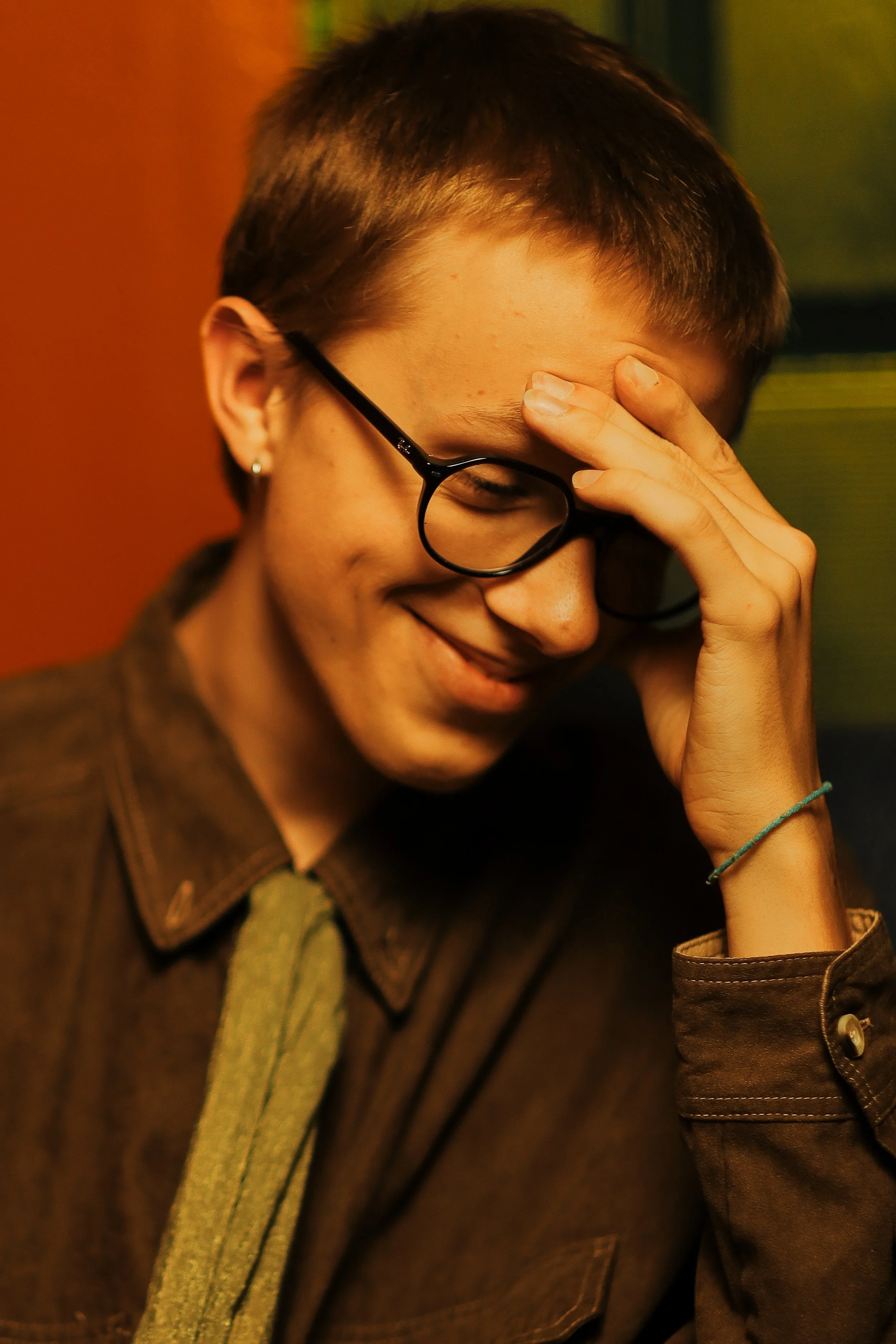 A person with short hair wearing glasses smiling with one hand on their forehead. They are dressed in a brown jacket and a greenish scarf, with an earring in one ear, and a colorful background.