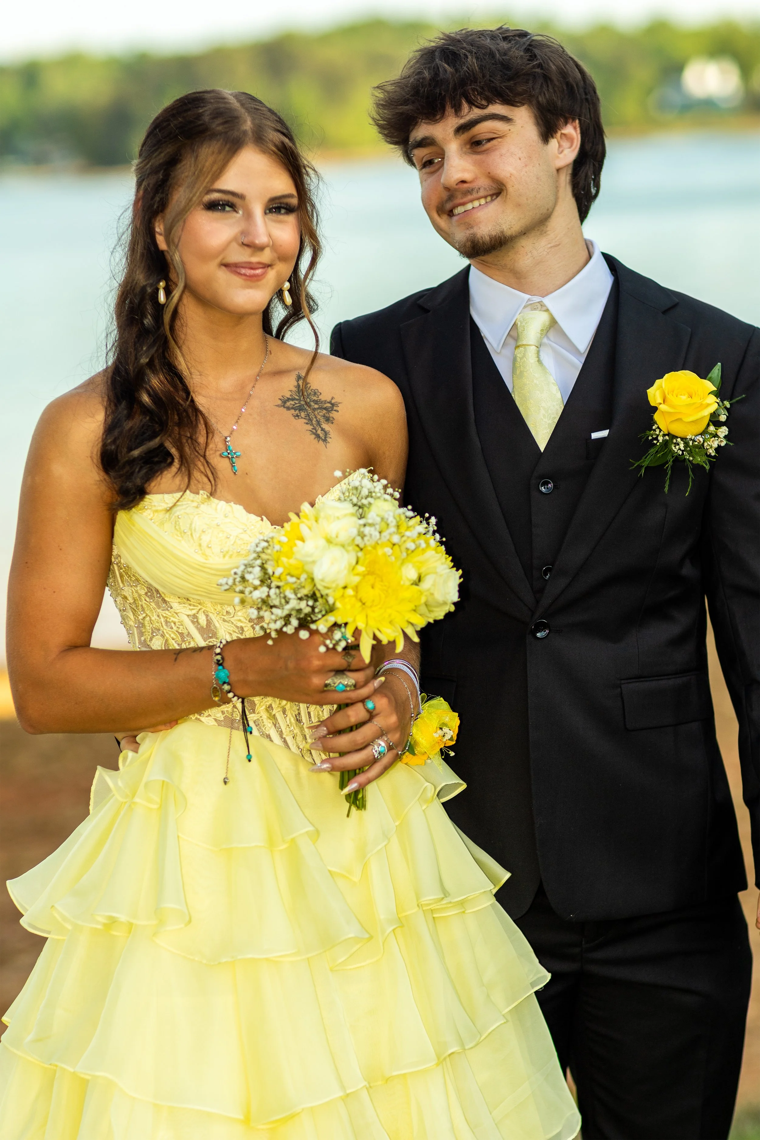 A young woman in a yellow dress holding a bouquet of yellow and white flowers, standing beside a young man in a black suit with a yellow boutonniere, outdoors near a body of water.