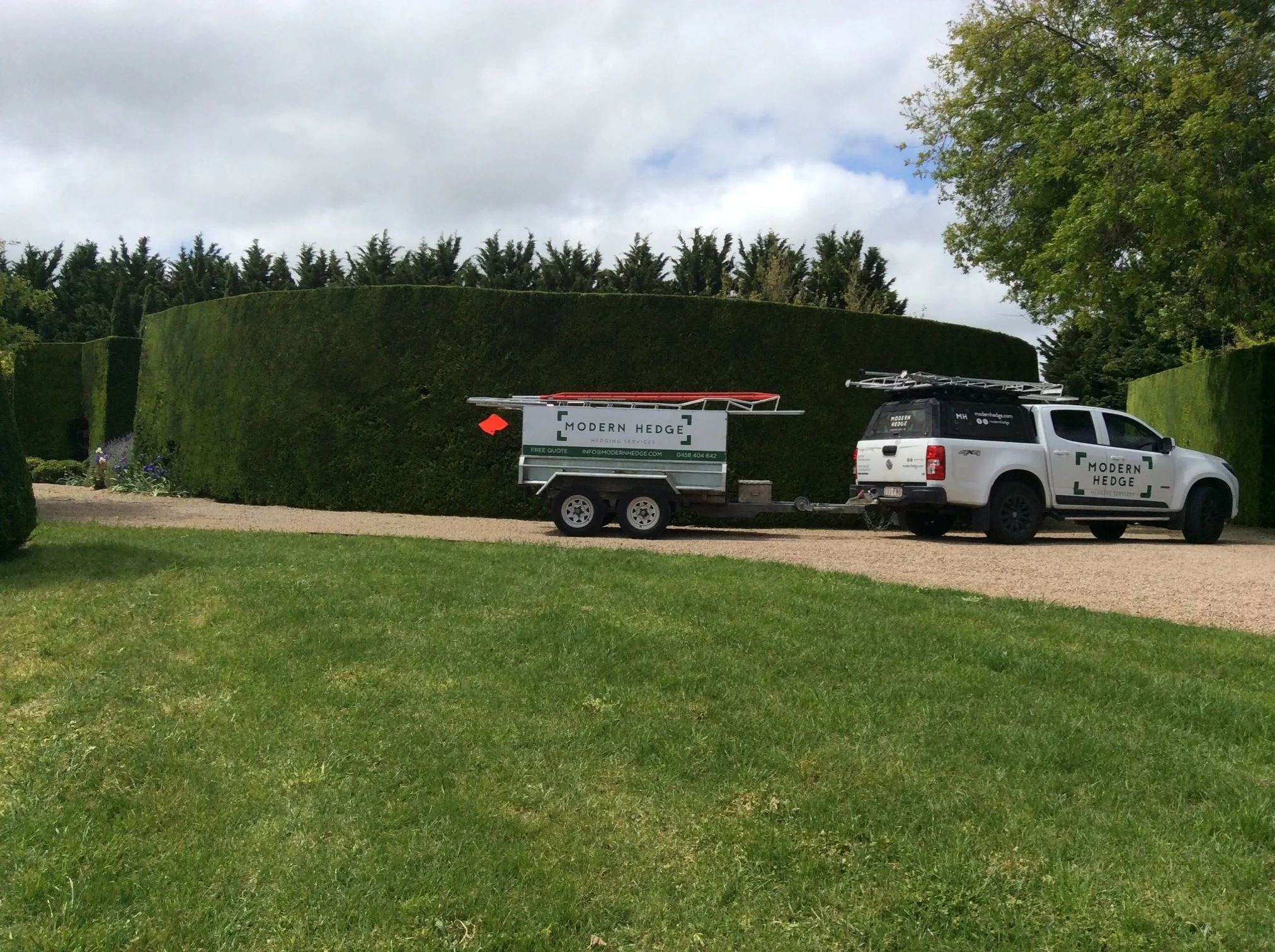 A 'Modern Hedge' branding parked on a gravel driveway, towing a trailer with trimming tools, in front of tall, neatly trimmed hedges and greenery.