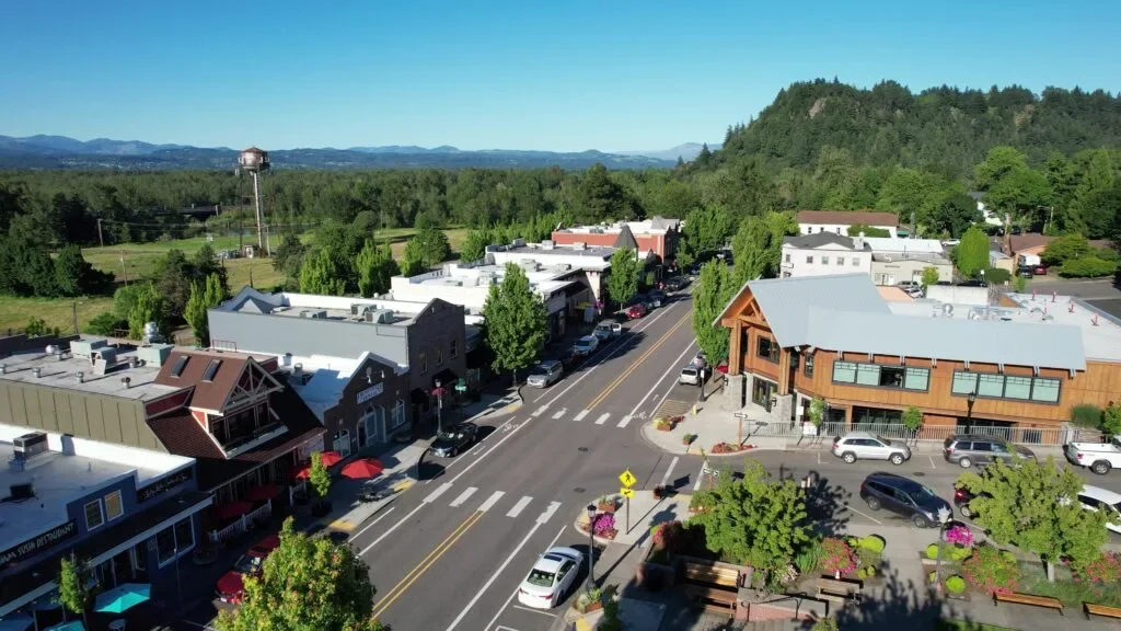 A small town main street with shops, parked cars, trees, and a mountain in the background under a clear blue sky. Town center of Troutdale, OR.