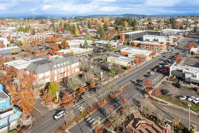 Aerial view of a small urban town with trees showing fall colors, streets, cars, and mixed commercial and residential buildings under a cloudy sky.