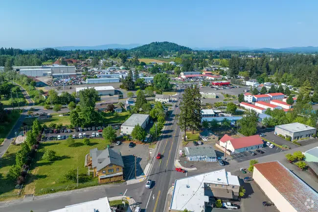 Aerial view of a small town with shops, houses, and trees, set against a backdrop of green hills and blue sky. City center of Boring, OR.