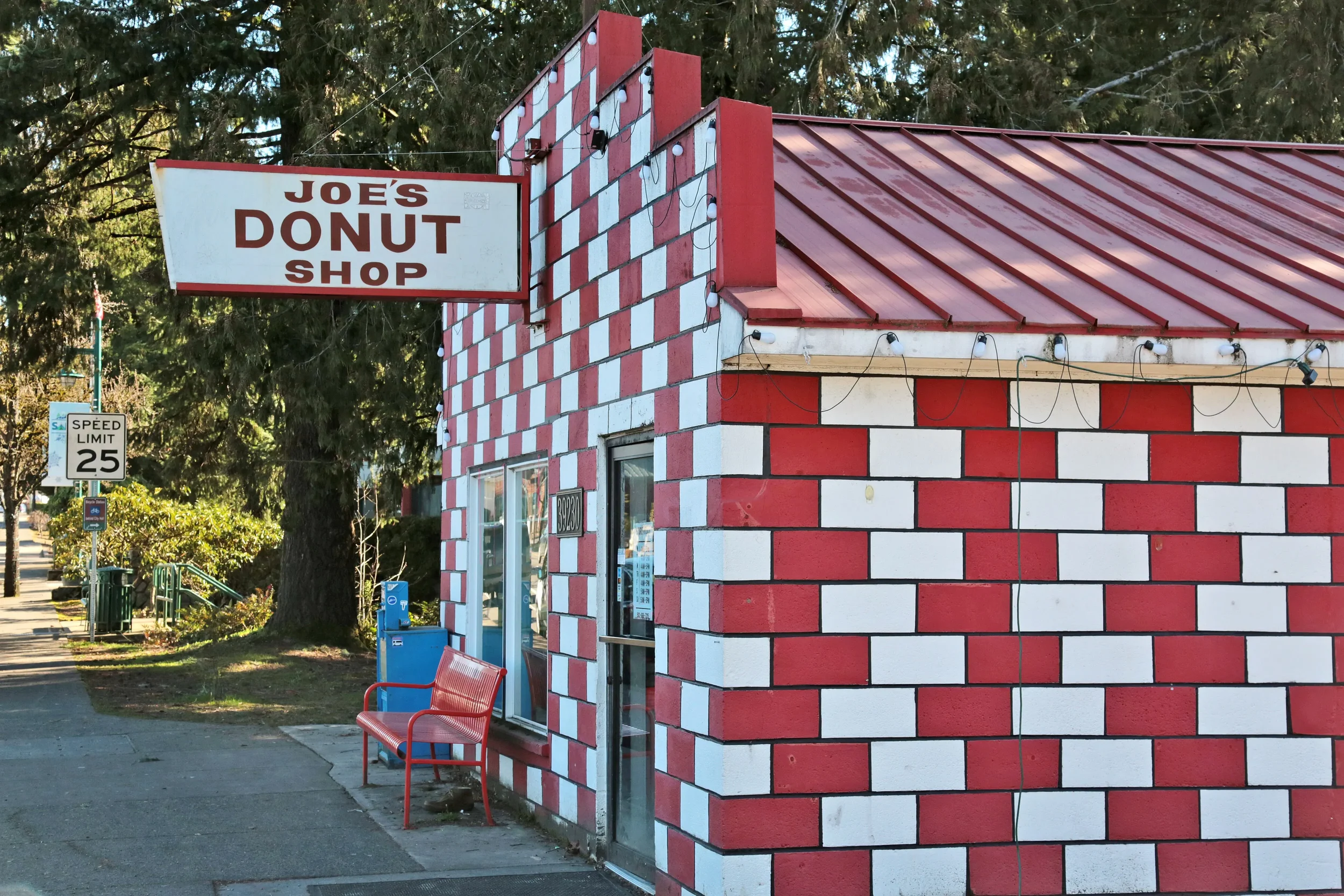 A red and white brick building with a red metal roof, large sign that reads 'Joe's Donut Shop,' string of lights along the roof, a red bench outside, and trees in the background. Sandy, OR.