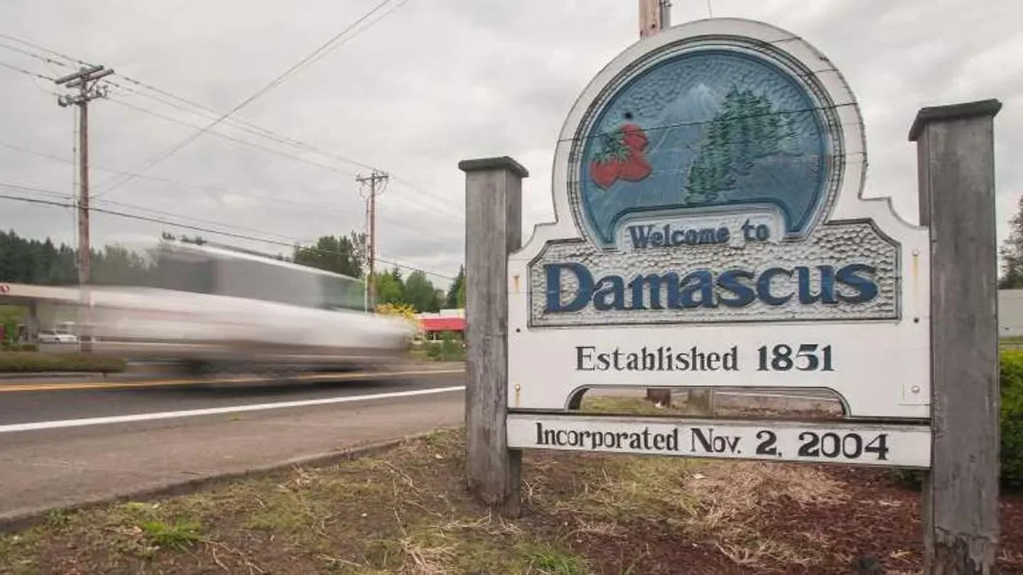 Welcome to Damascus sign with established date 1851 and incorporation date November 2, 2004, next to a road with a blurred vehicle passing by and power lines overhead. Damascus, OR.