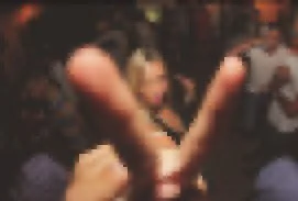 Imagen borrosa de un grupo de personas en una reunión social, con énfasis en una mano en primer plano haciendo una señal de corazón con los dedos.