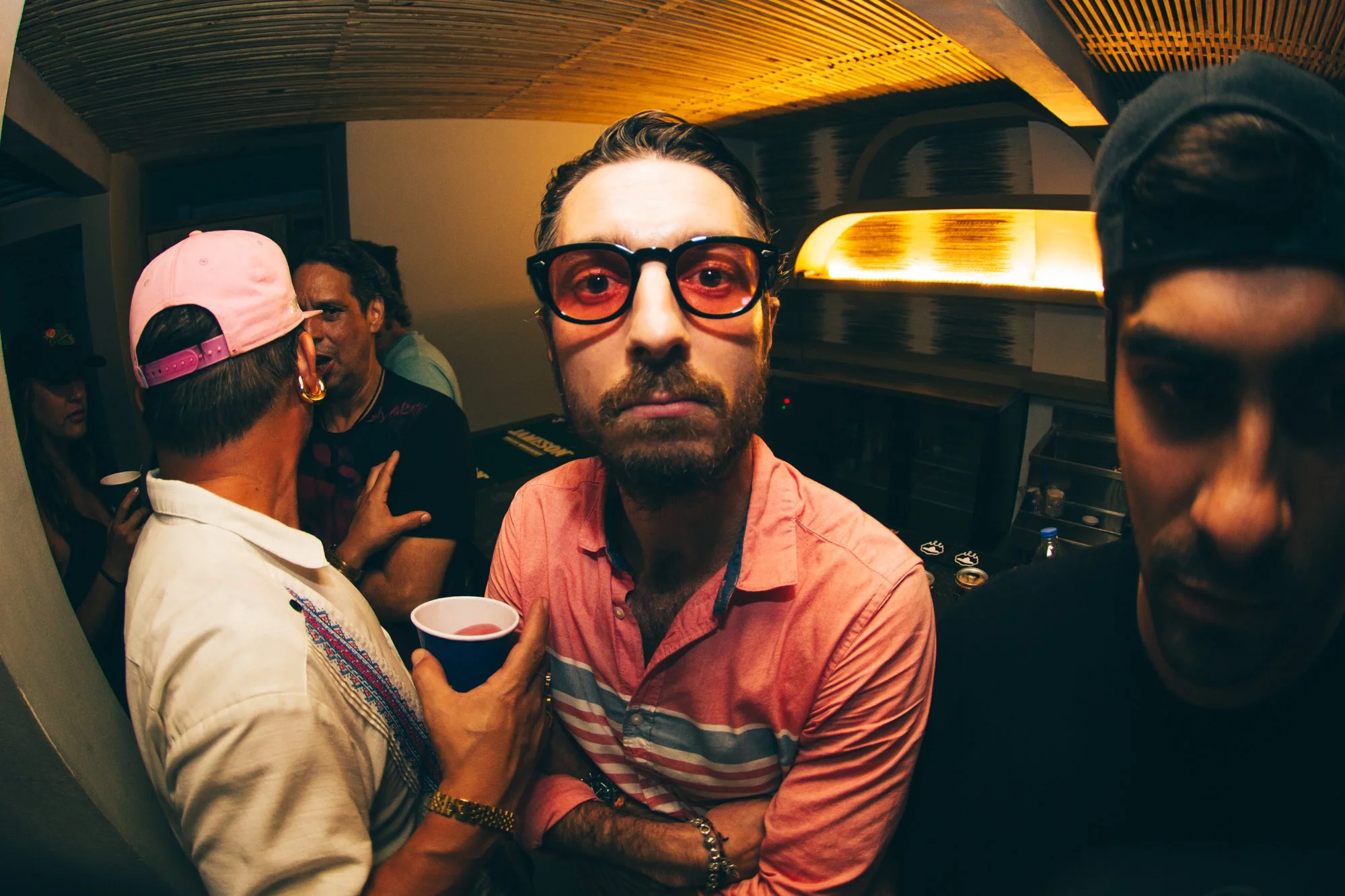Grupo de personas en una fiesta, un hombre en el centro lleva gafas rojas y camiseta rosa, una mujer a la izquierda con gorra rosa y camisa blanca sostiene un vaso con bebida, otra persona en la esquina derecha con gorra oscura. Ambiente con luz cálida y moderna.