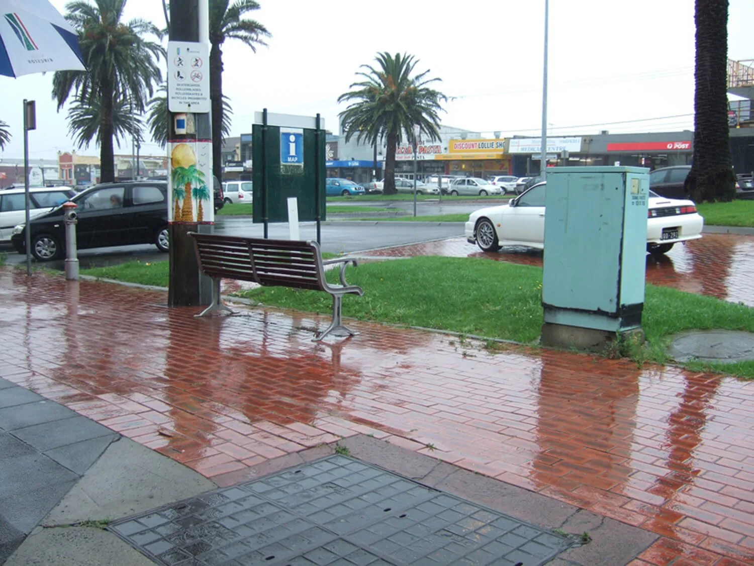 A rainy day scene at a bus stop with a wet tiled sidewalk, a bench, a large palm tree, and parked cars, with a row of stores in the background.