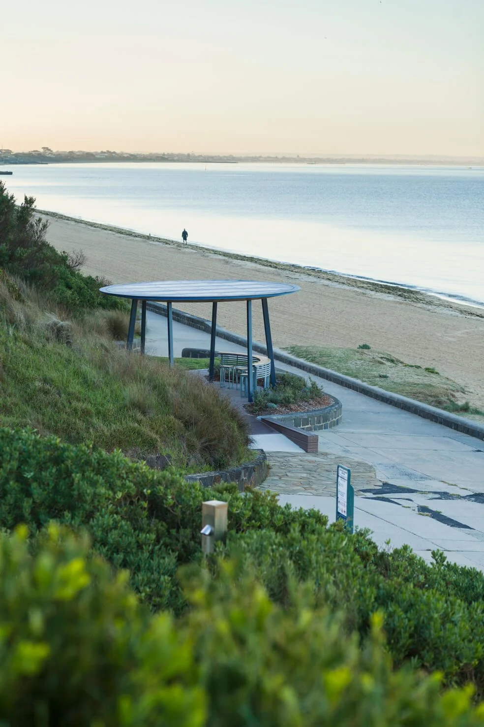 A peaceful beach scene with a sandy shoreline, calm water, a person walking along the beach, and a covered seating area with a round roof on a paved pathway.