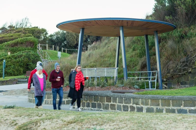 Four people walking in a park, with a shelter that has a metal bench underneath it, surrounded by grass and shrubbery.