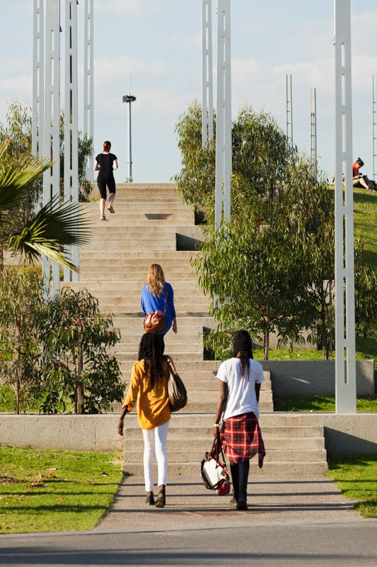 Three women walking up outdoor concrete stairs toward an open park area with other people sitting and walking around, surrounded by green trees and plants, under a partly cloudy sky.
