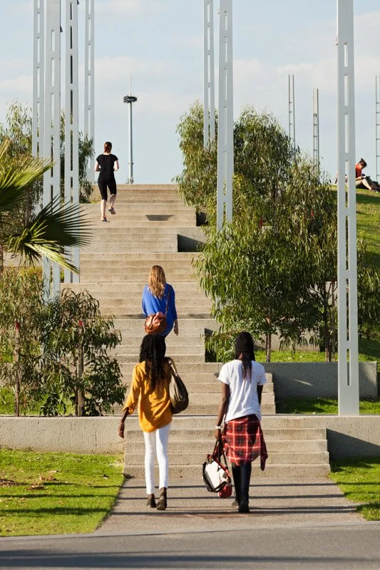 Three women walking up outdoor concrete stairs toward an open park area with other people sitting and walking around, surrounded by green trees and plants, under a partly cloudy sky.