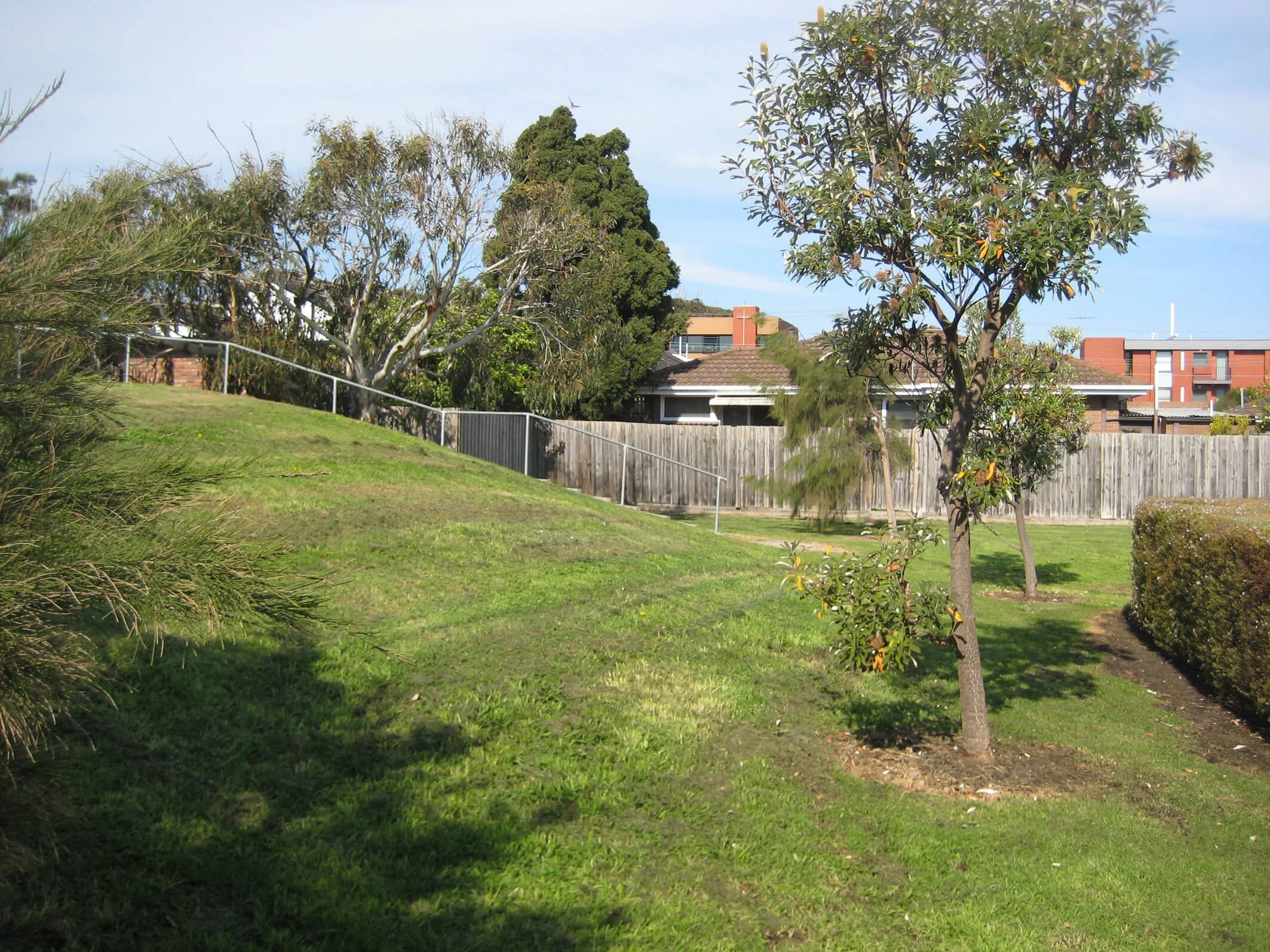 A grassy park with trees, a wooden fence, and residential buildings in the background under a clear blue sky.