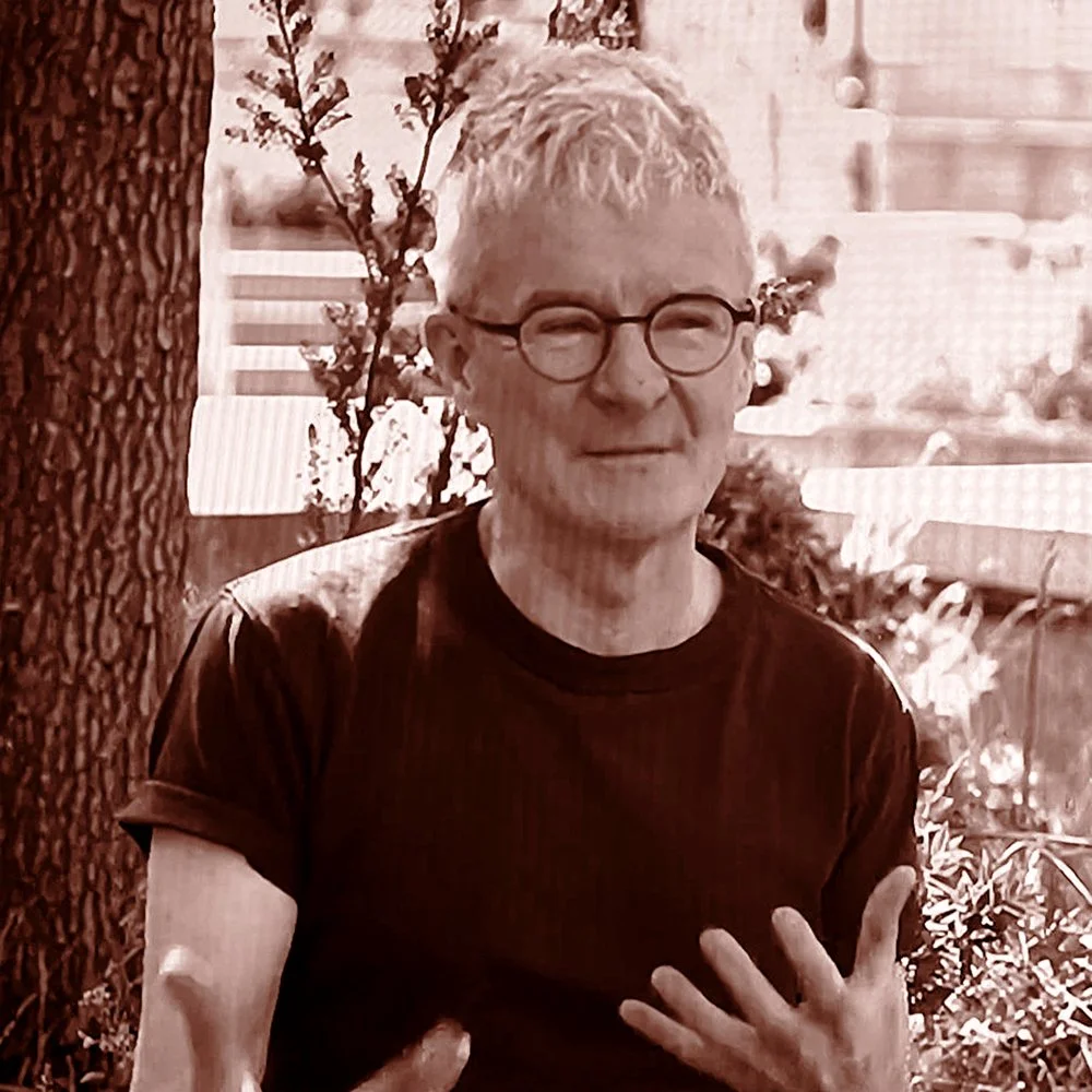 A man with light hair, round glasses, wearing a dark t-shirt, standing outdoors near a tree and plants, with a house in the background.