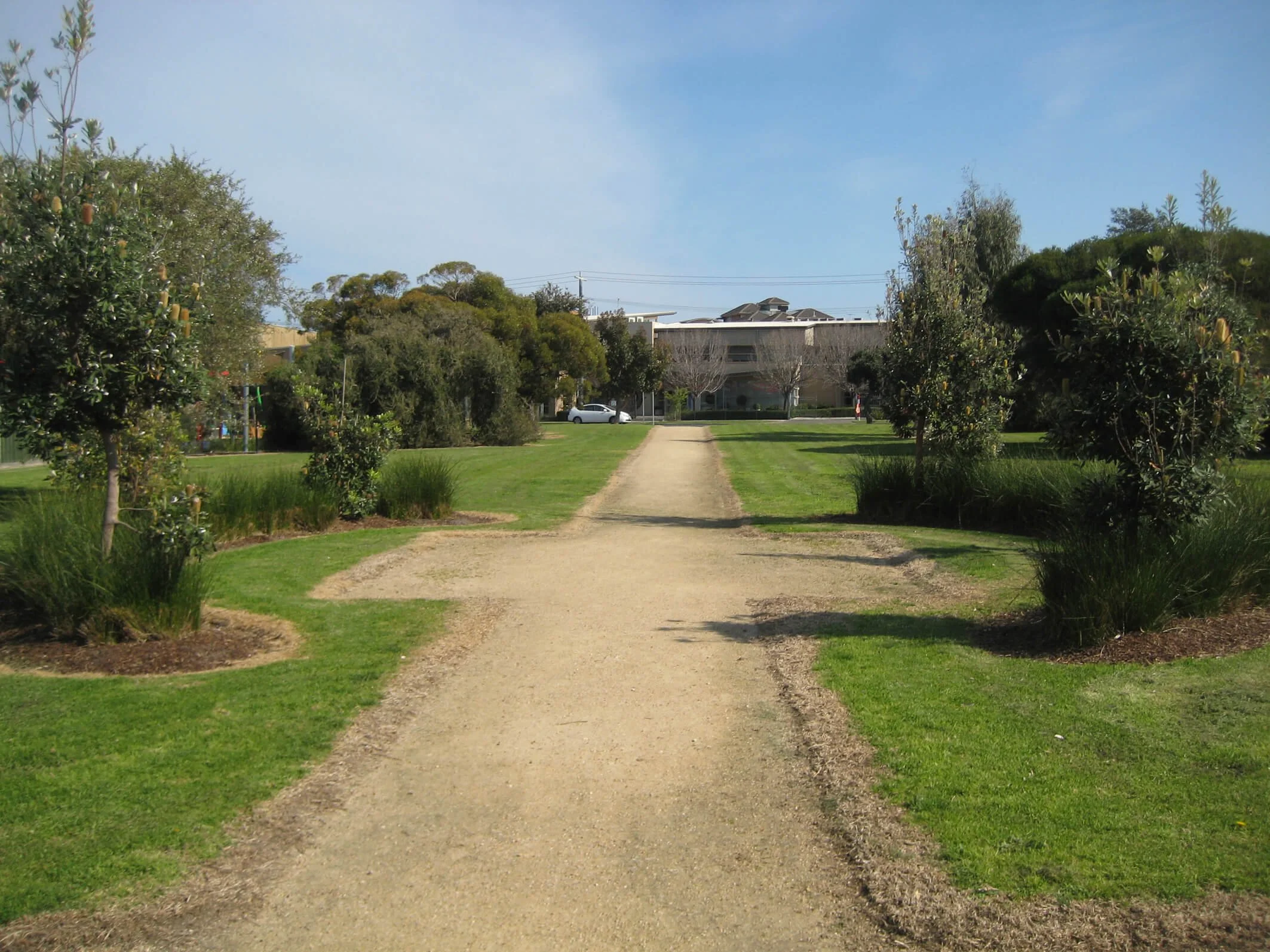 A park with a dirt pathway running through green grass and trees on both sides, under a partly cloudy sky.
