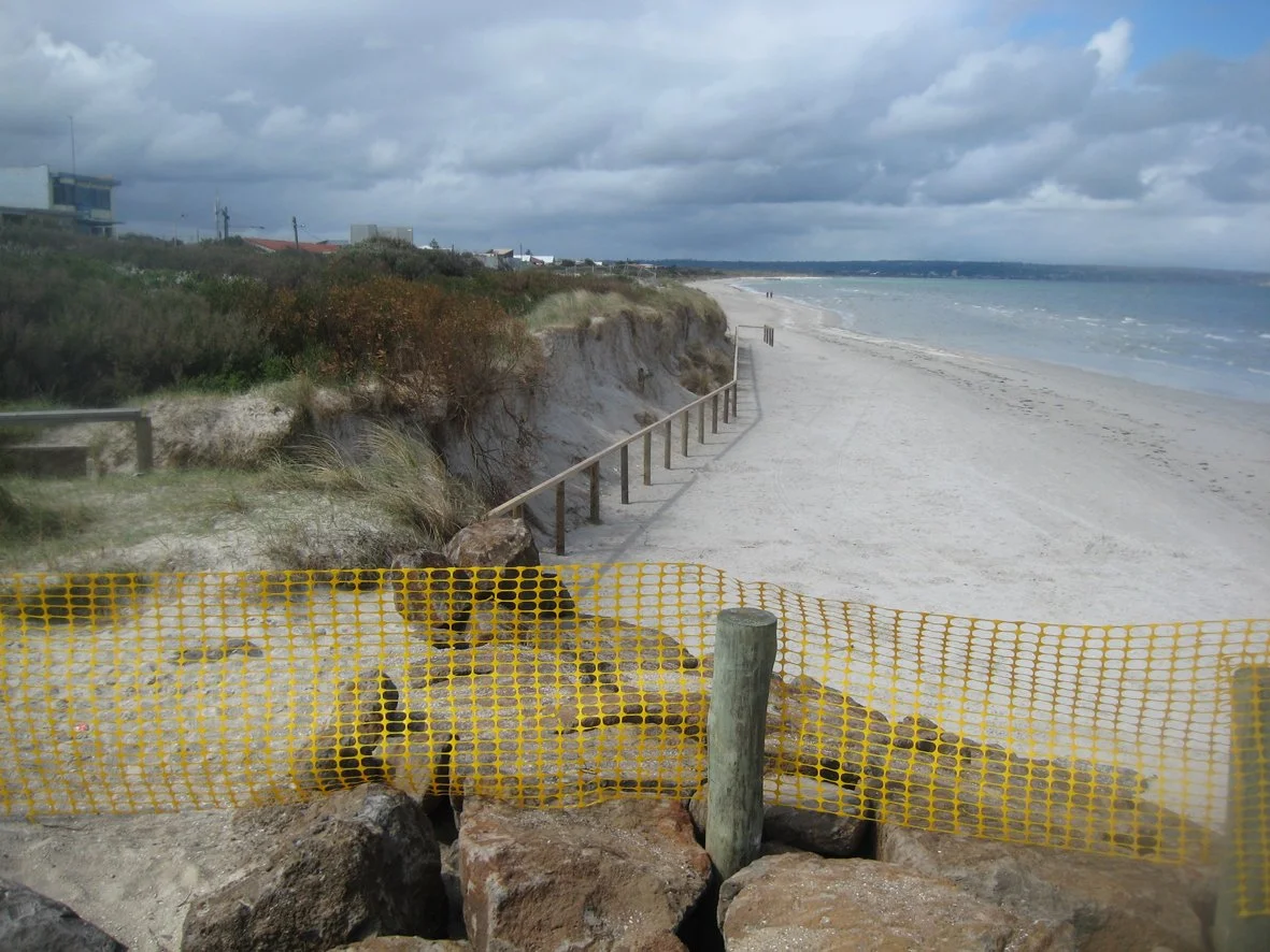 A beach with a partially blocked access point marked by yellow construction barricade, overlooking a sandy shoreline with a cliff and ocean in the background under cloudy skies.