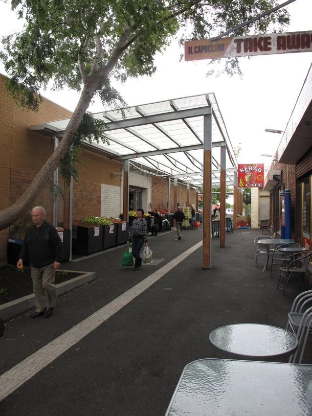 An outdoor marketplace with manual produce stands and people shopping on a sidewalk with a canopy overhead and a sign for kebab and cafe in the background.