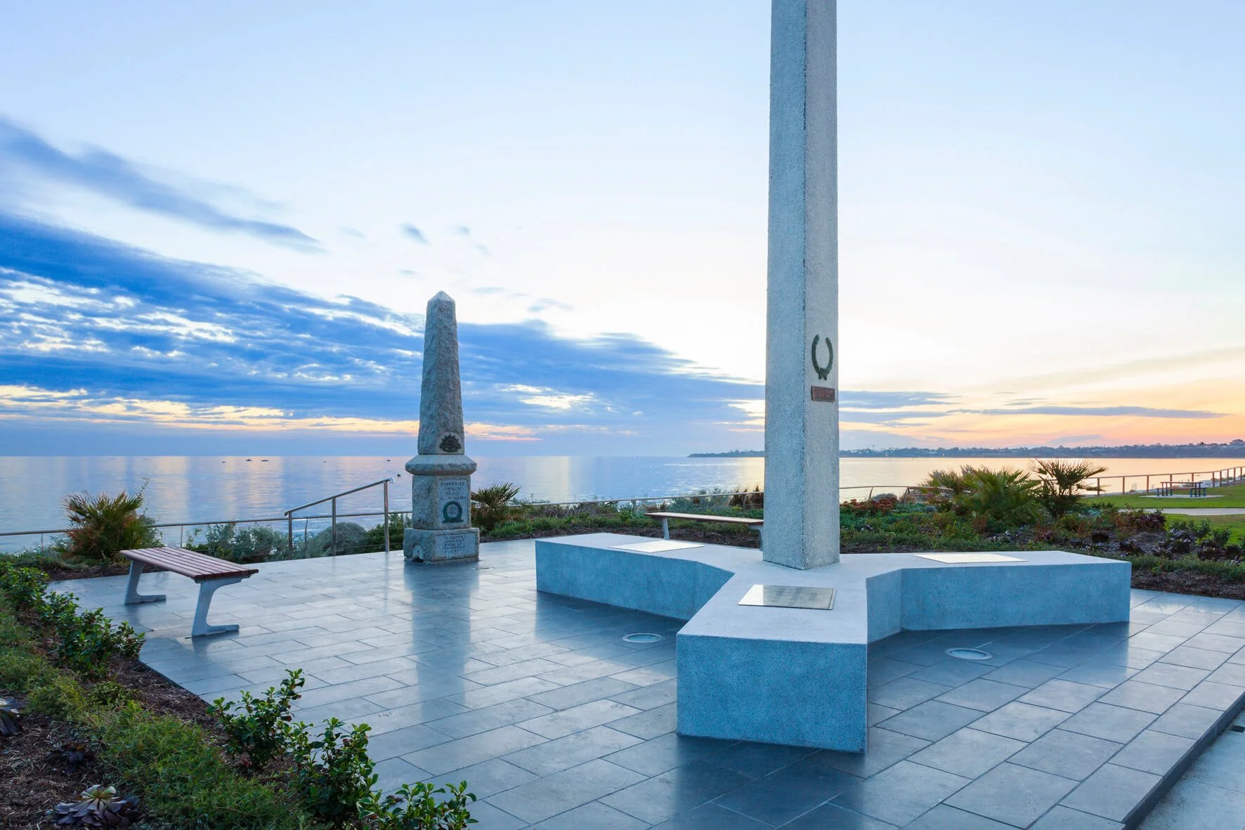 Memorial monument with obelisk, horseshoe-shaped column, and plaques, overlooking a body of water at sunrise, with benches and landscaping nearby.