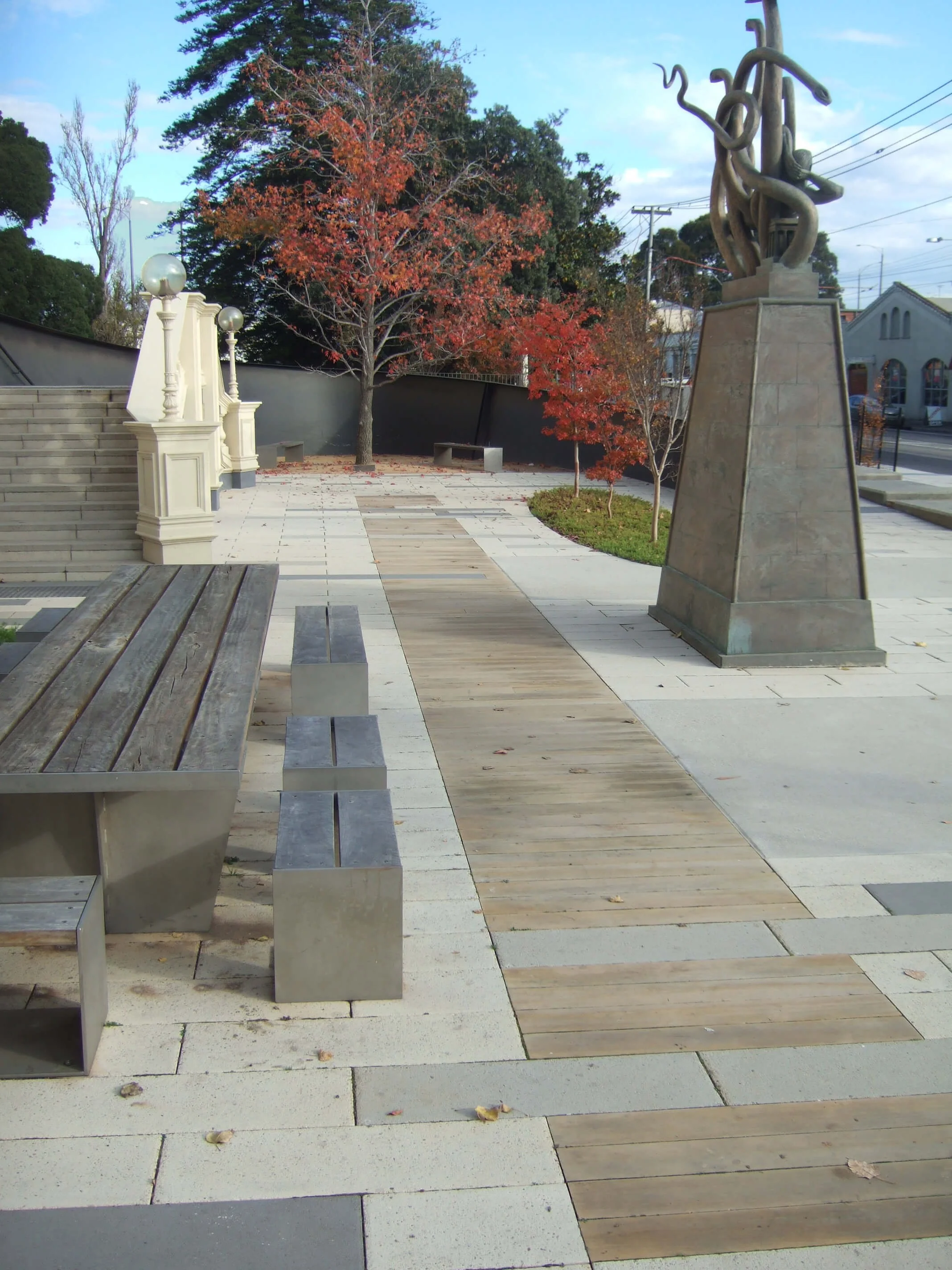 Public park or plaza with benches, a paved walkway, leafless trees with red and orange leaves, a sculpture on a pedestal, and a staircase with decorative lamp posts.