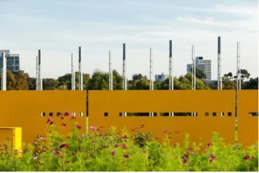 A yellow fence with small square and rectangular cutouts, with pink flowers and greenery in the foreground, and a city skyline with trees and tall buildings in the background under a partly cloudy sky.