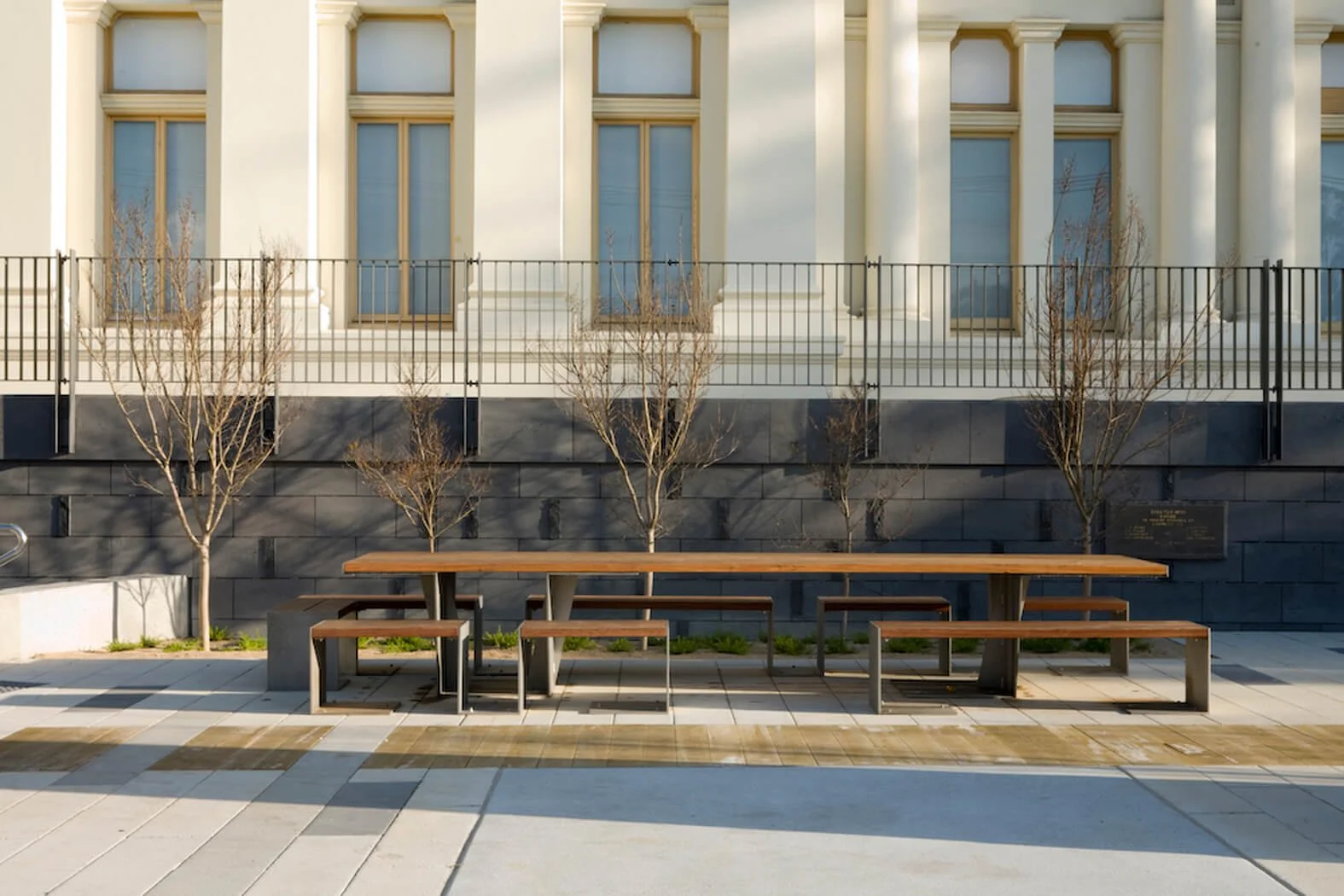 Empty outdoor seating area with wooden tables and benches, small leafless trees, a black metal fence, and a building with large windows in the background.