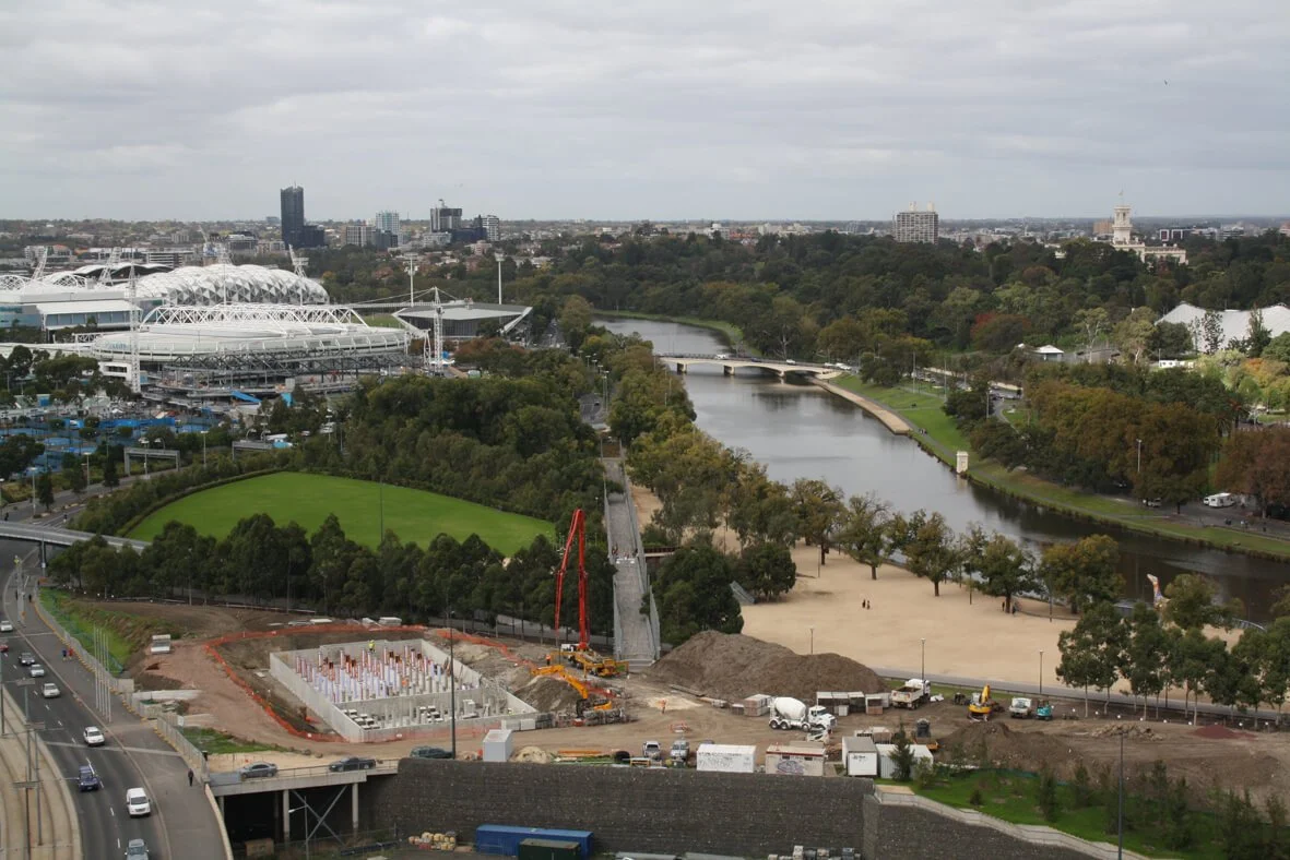 A cityscape showing a river running through a park with trees, a sports stadium, and residential buildings in the background. Construction equipment and a foundation are visible in the foreground.