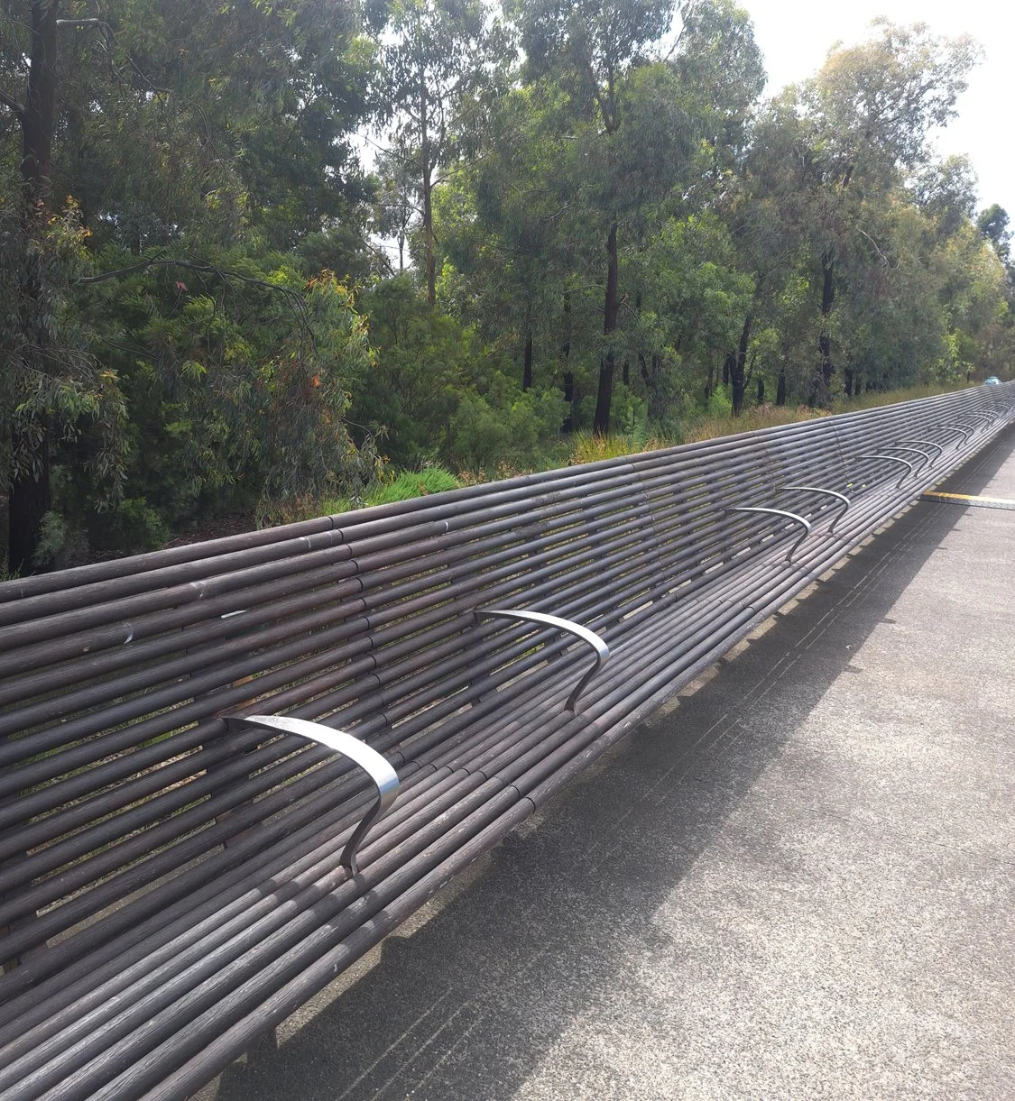 A long, curved metal bench with armrests on a sidewalk next to a forested area.