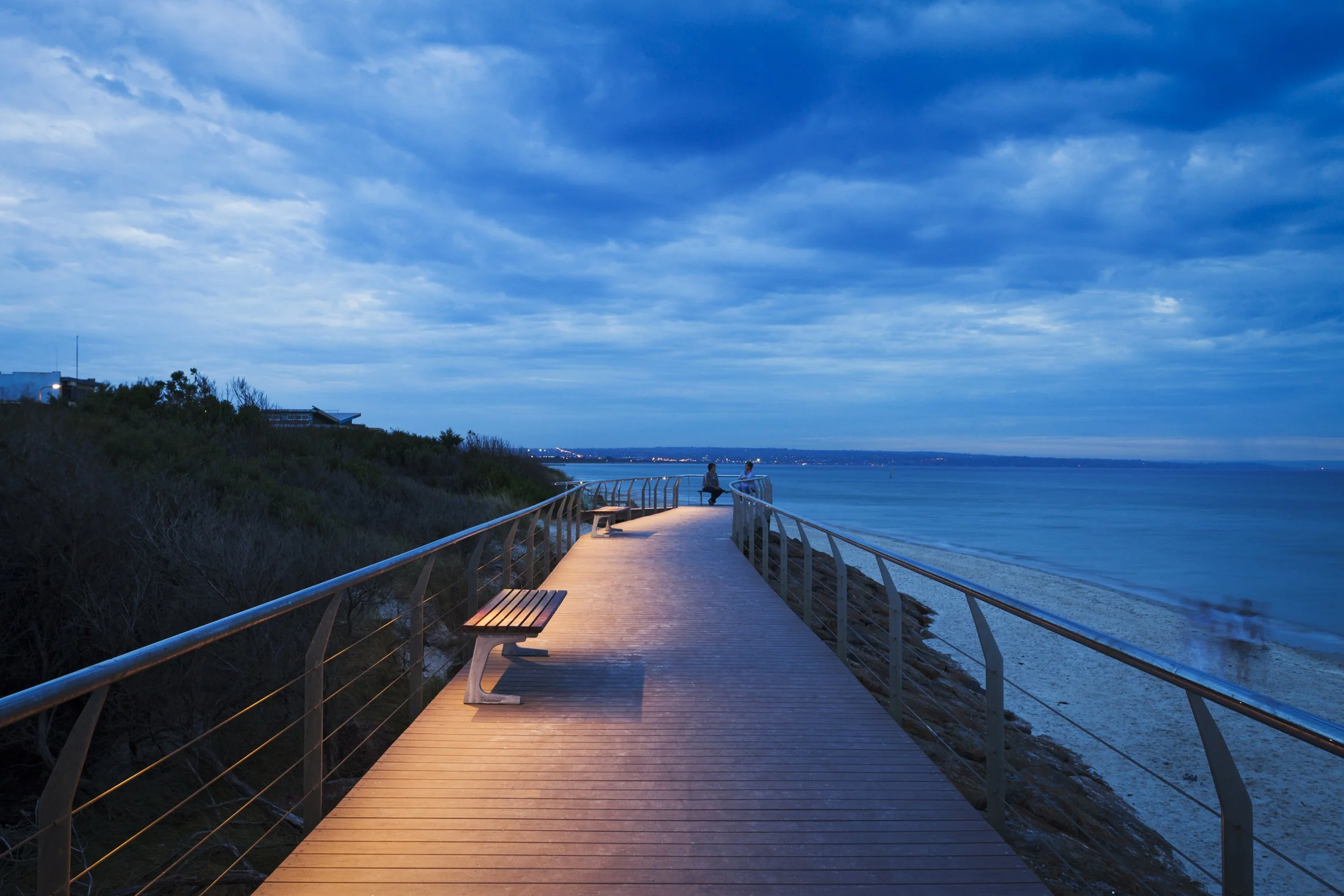 A wooden pier extending over the ocean at dusk, with two people sitting at the end and several benches along the walkway, under a partly cloudy sky.