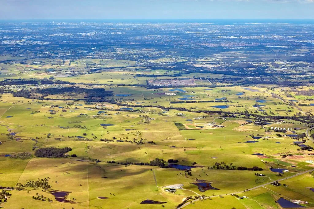 Aerial view of a rural landscape with green fields, small lakes, and scattered buildings, under a partly cloudy sky.