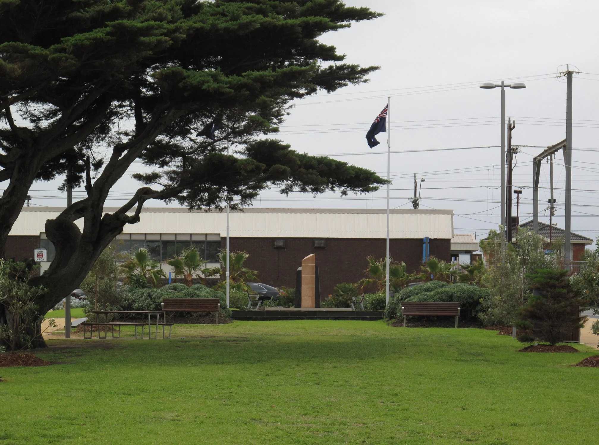 Park with a large tree, green grass, benches, and a flagpole with the Australian flag, across from a building with bushes and palm trees.