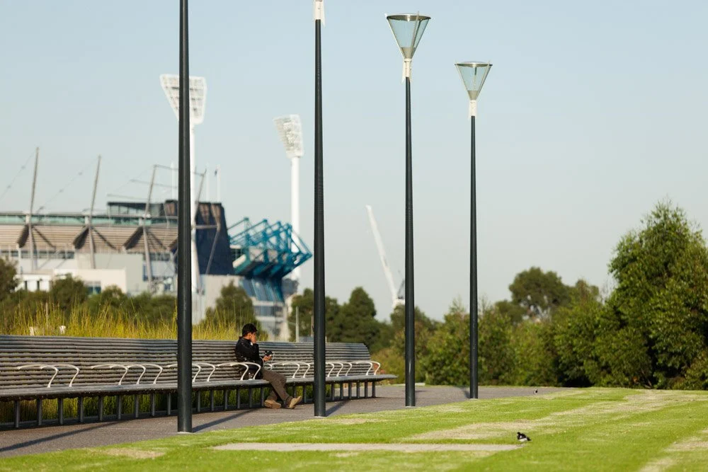 A person sitting on a bench in a park, looking at a cell phone, with four tall streetlights and a stadium or arena in the background, along with green trees and a bird on the grass.