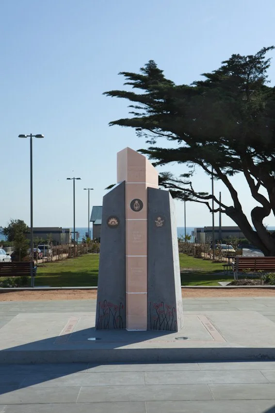 A memorial monument with plaques, situated in a park with benches, trees, and a view of the ocean in the background.