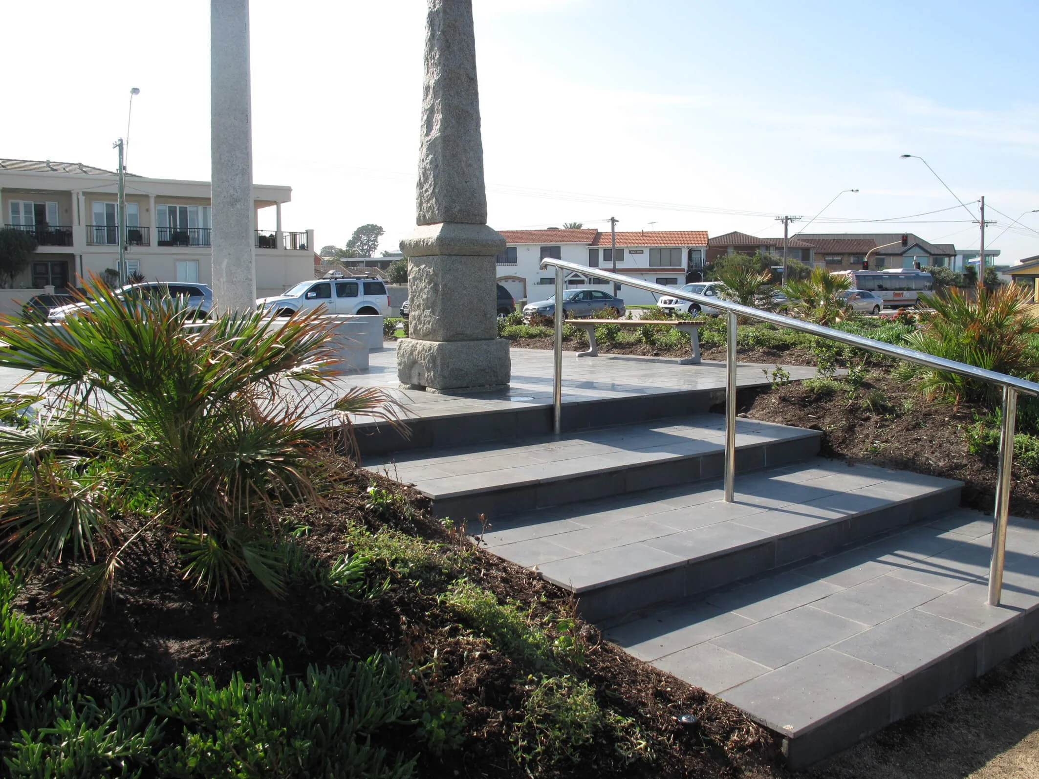 Concrete steps with metal handrail leading up to a platform with a stone monument, bicycle, and benches, surrounded by landscaped greenery, with residential buildings and parked cars in the background.